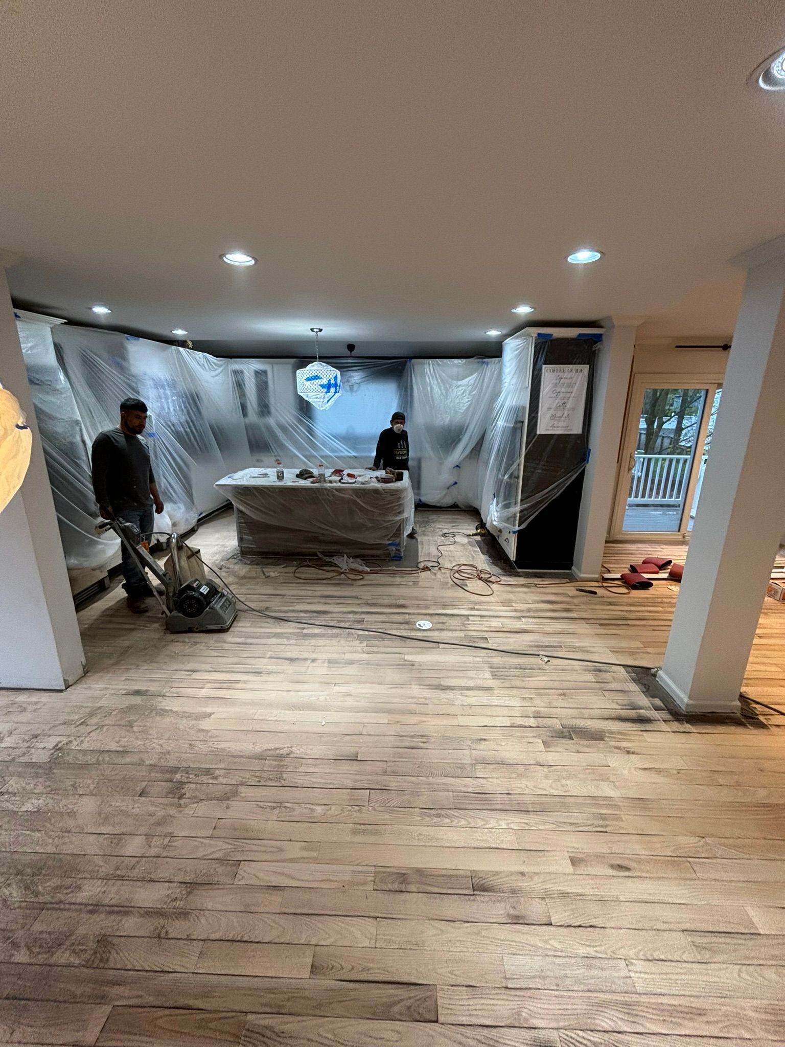 Workers sanding a wooden floor in a room. Cabinets and appliances are covered in plastic sheeting.