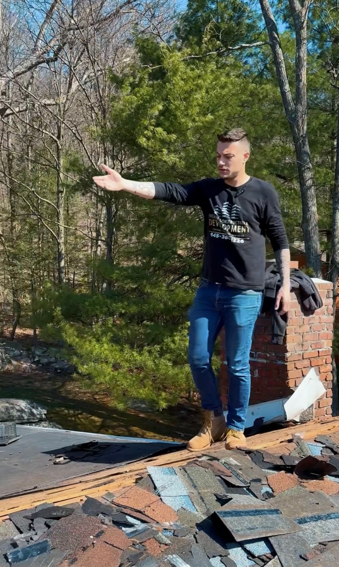 Man on a roof pointing to the side, wearing jeans and work boots. Chimney and discarded shingles are nearby.