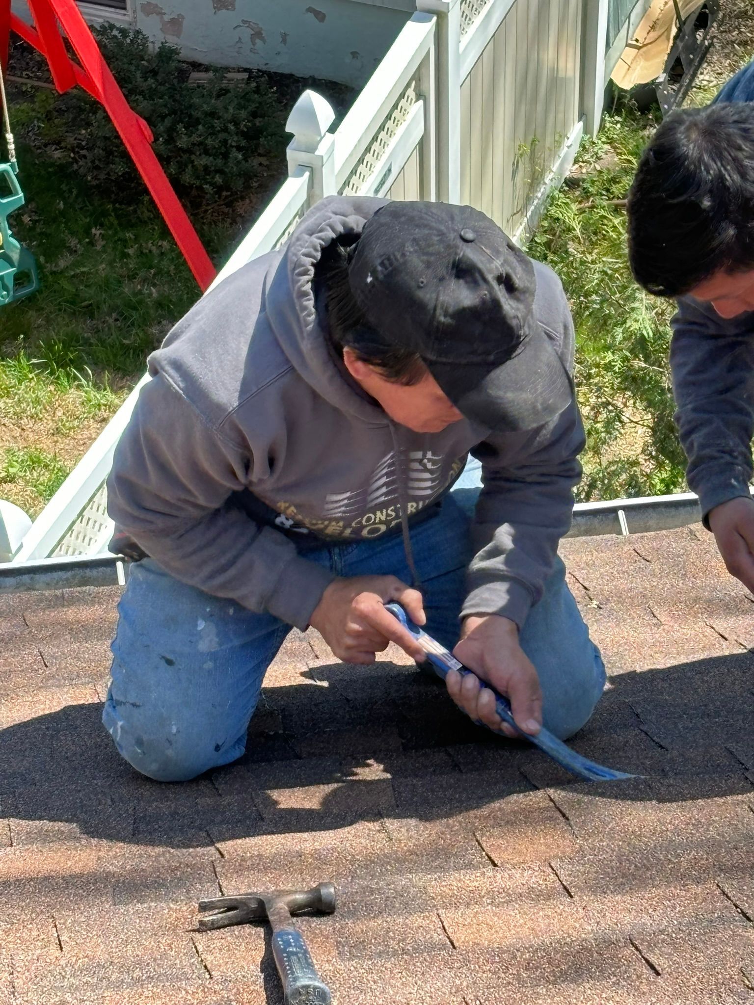 Person on a rooftop kneeling, using a tool to work on shingles.  A hammer is visible on the roof.
