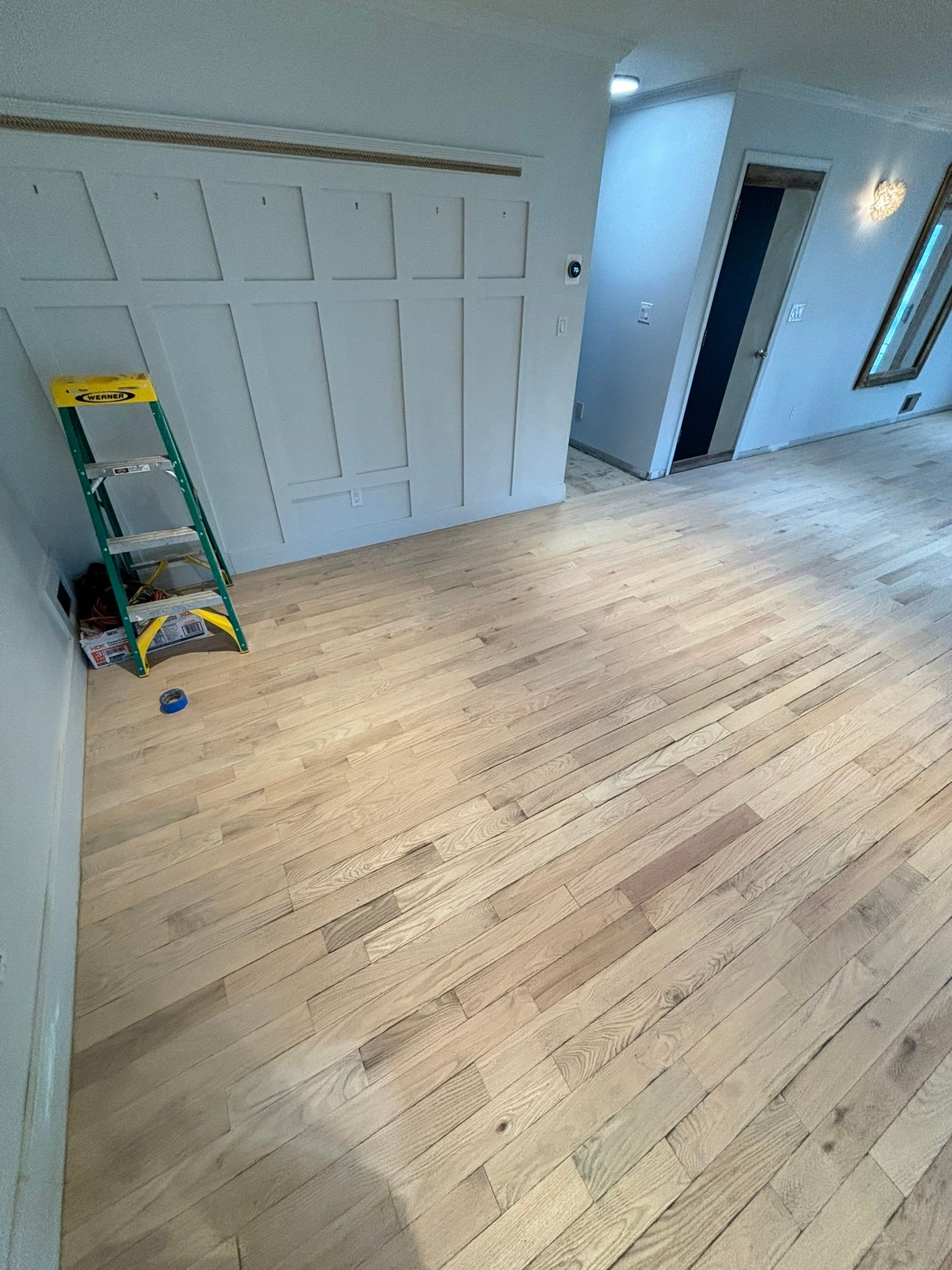 Light wood floor and wall with paneling, a green ladder, and an open doorway.