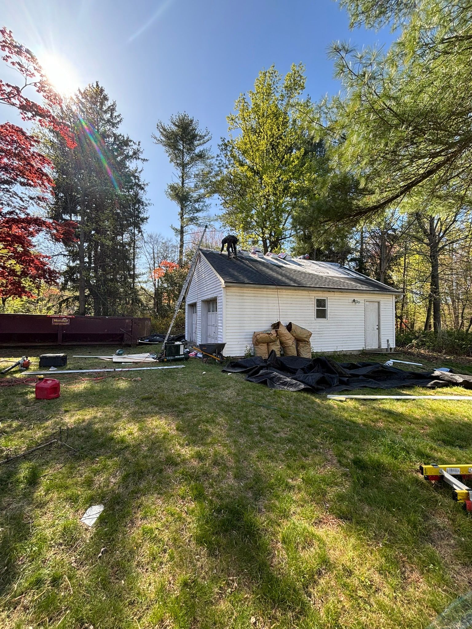 A white shed with a damaged roof surrounded by debris in a grassy yard under a sunny sky.
