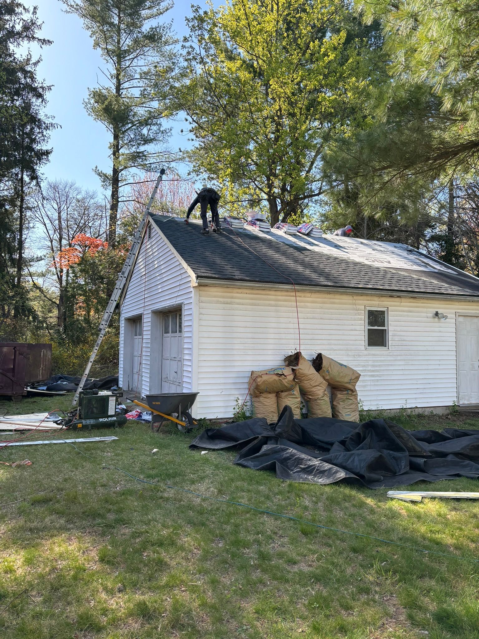 A person removing shingles from a white garage roof; ladder propped against the roof.