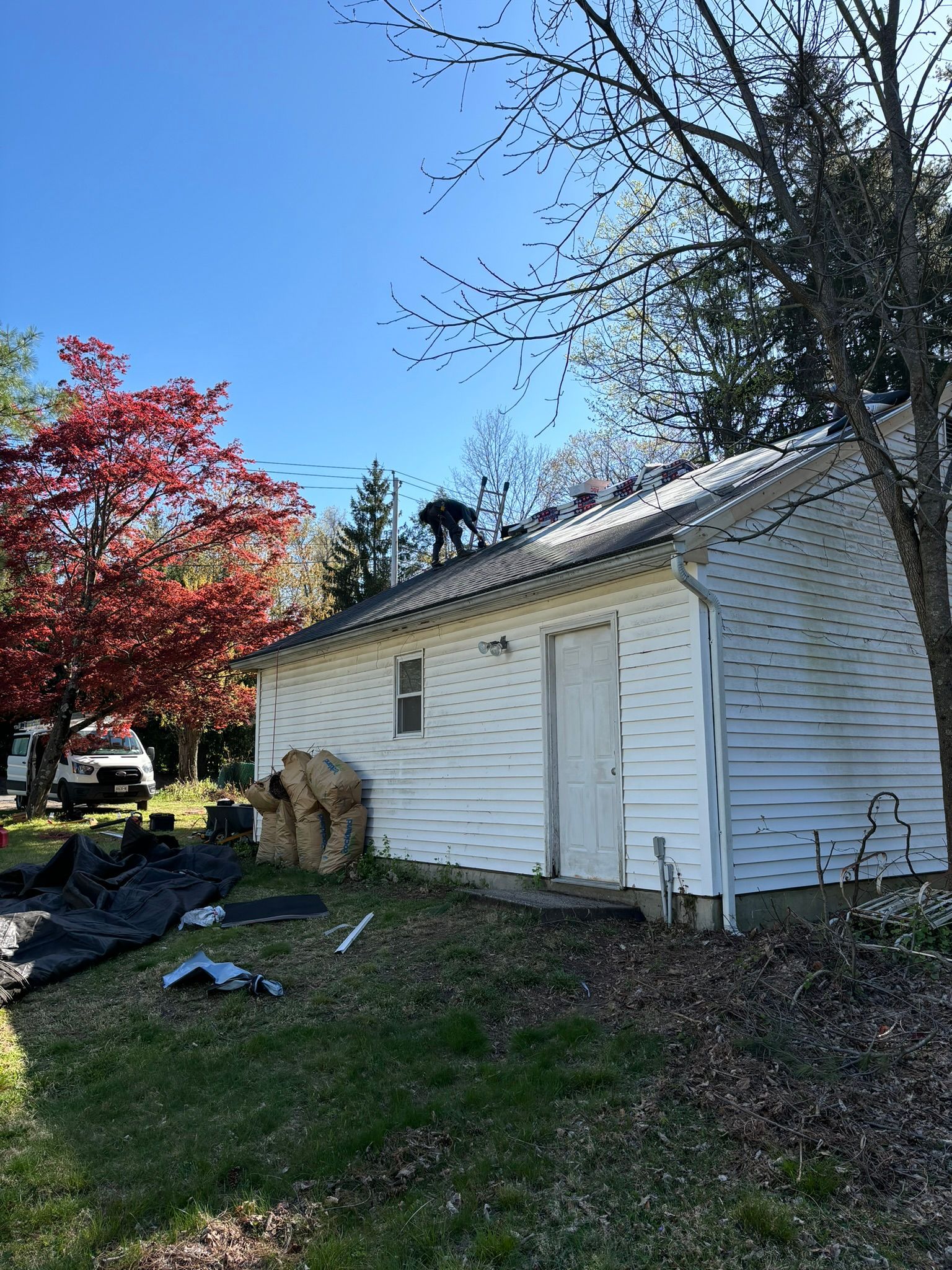 Person on a rooftop of a white house with debris on ground. Bright red and green trees in the background under a blue sky.