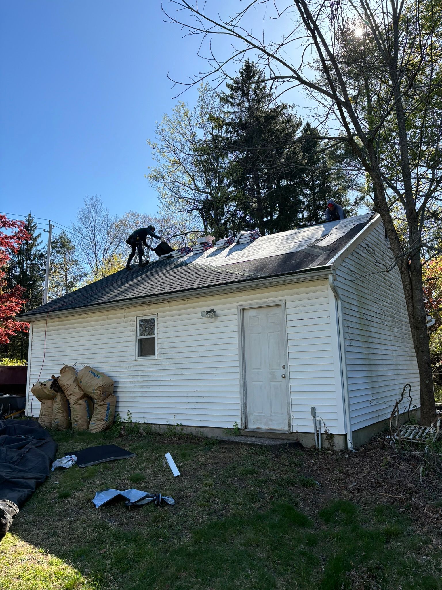 Workers on a roof, removing shingles from a white, small building under a blue sky.