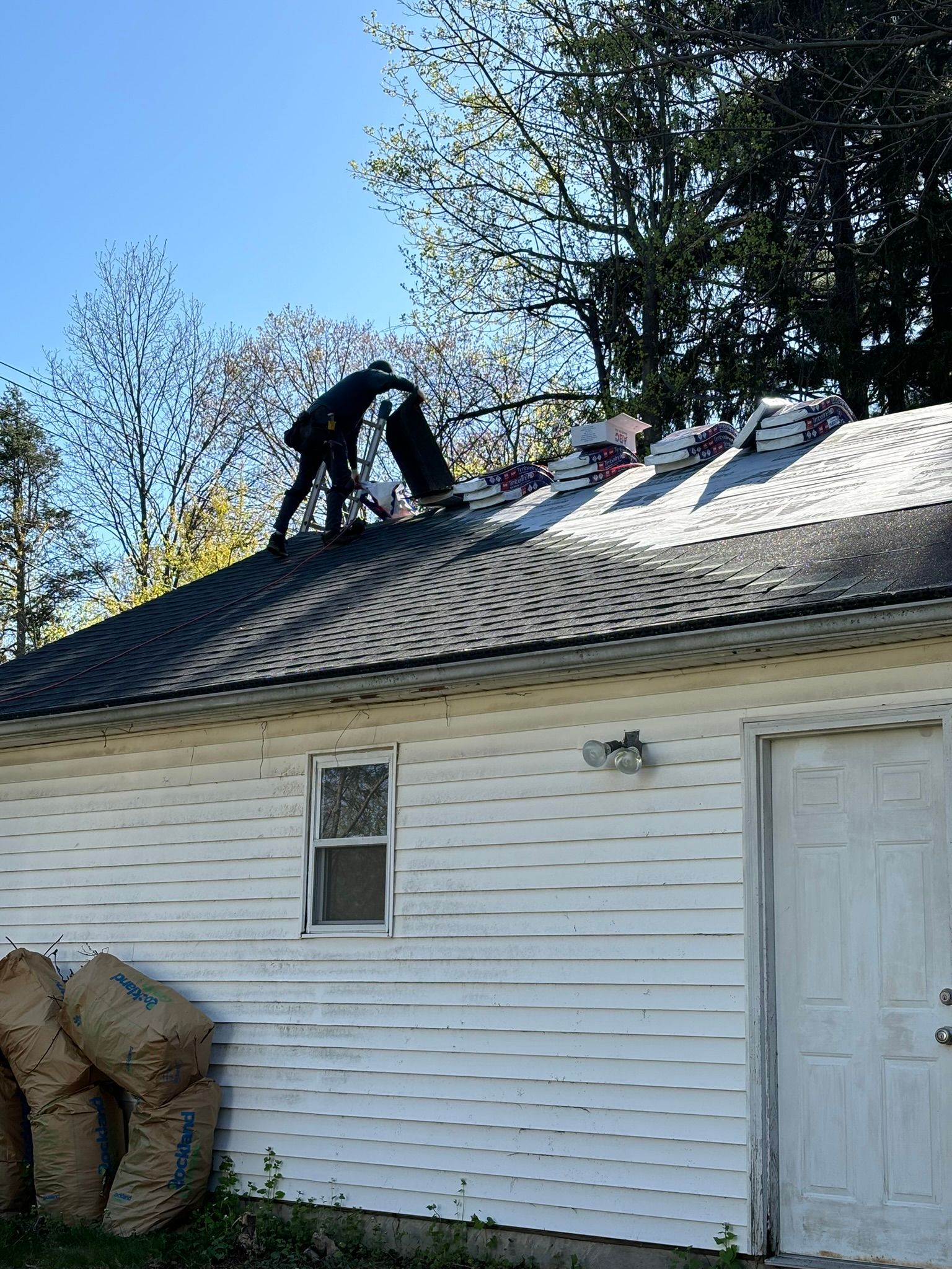 Person working on a roof, shingles visible. White building, blue sky, and foliage in background.