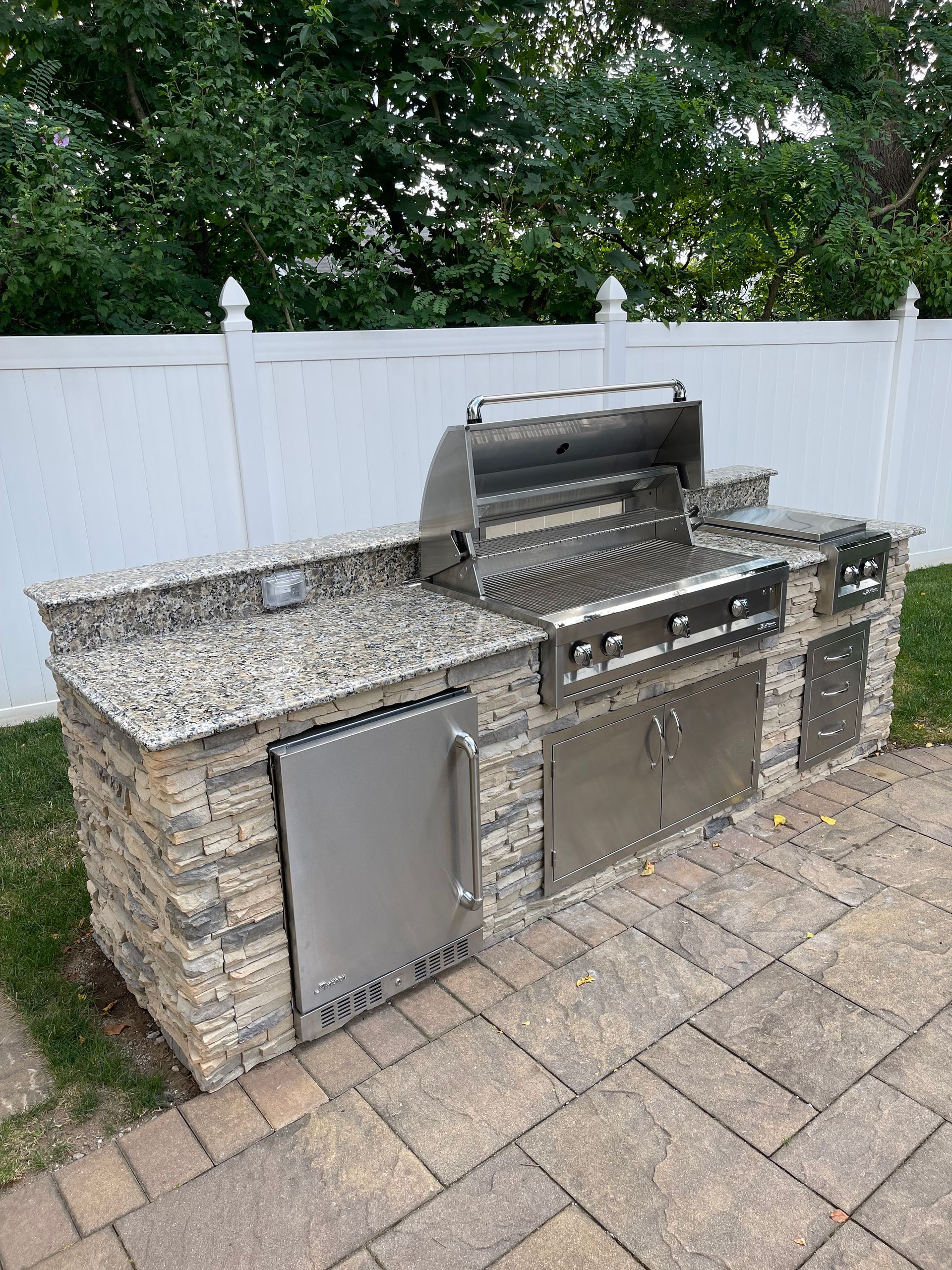 Outdoor kitchen with stainless steel grill, refrigerator, granite countertops, and stone veneer base.