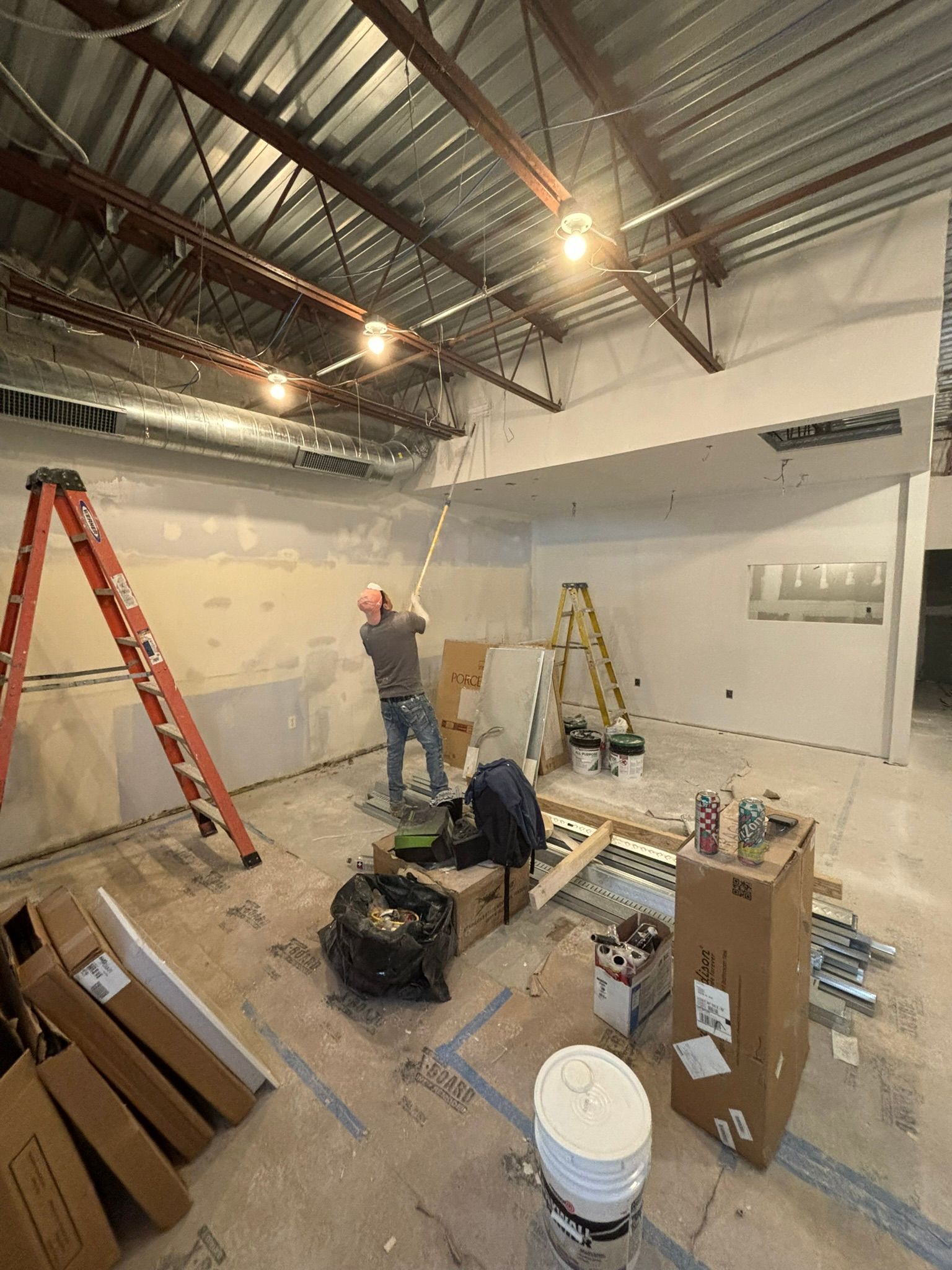 Construction in progress: a person on a ladder, painting a white wall. The room has exposed ceiling and drywall.