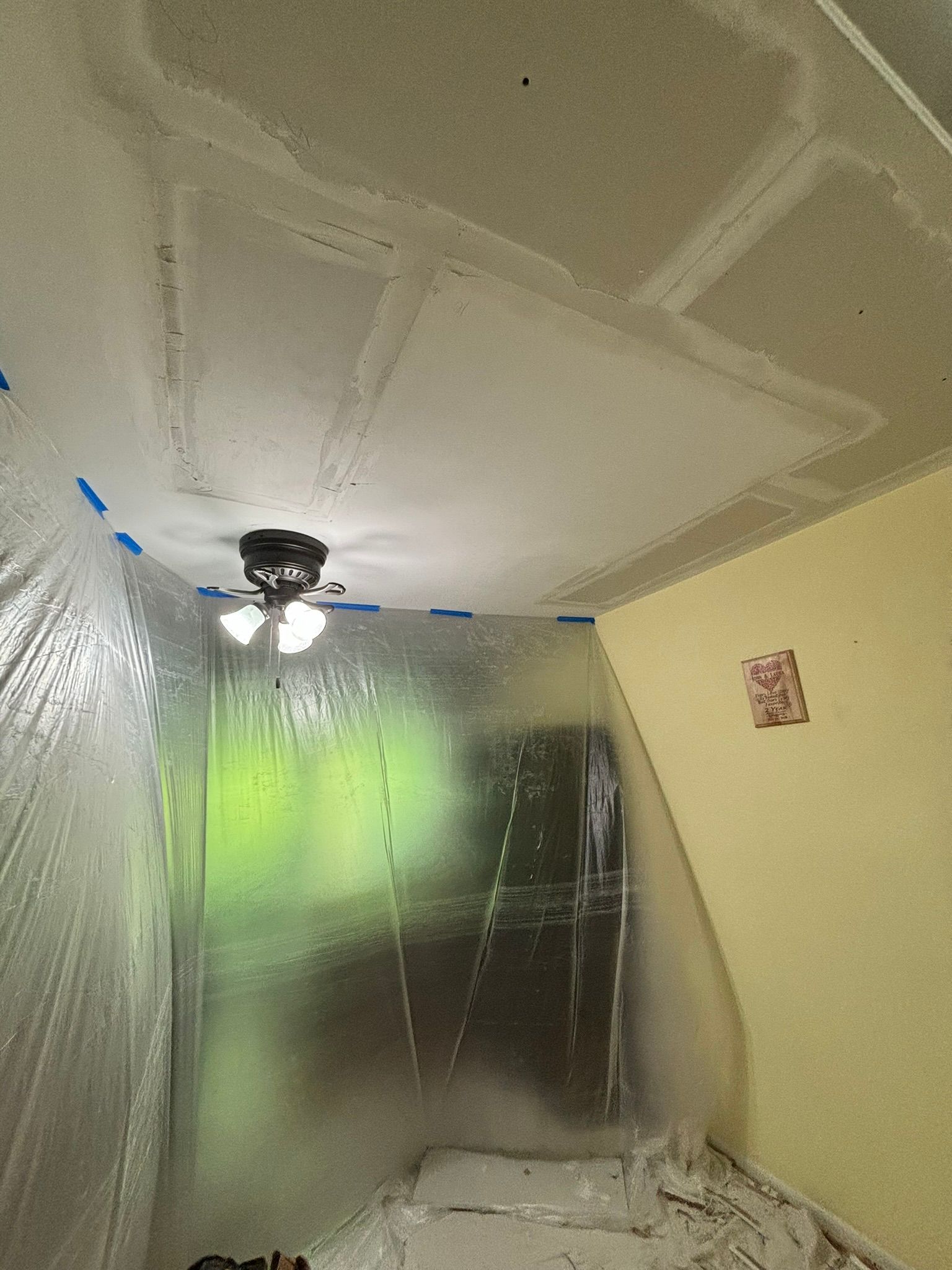 Room with plastic sheeting, ceiling repair in progress, partially lit by a ceiling fan.