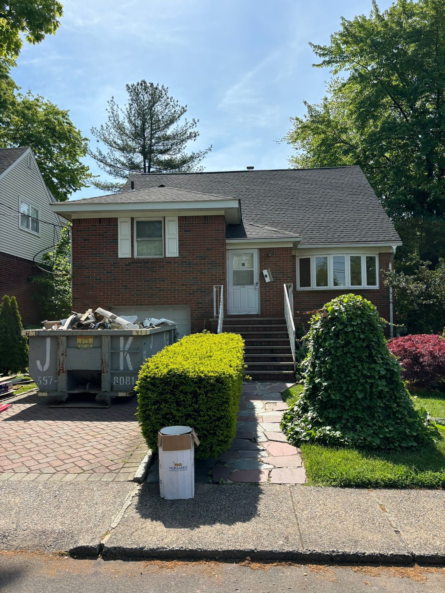 Brick house with a dumpster in front; overgrown bushes and a brick walkway.
