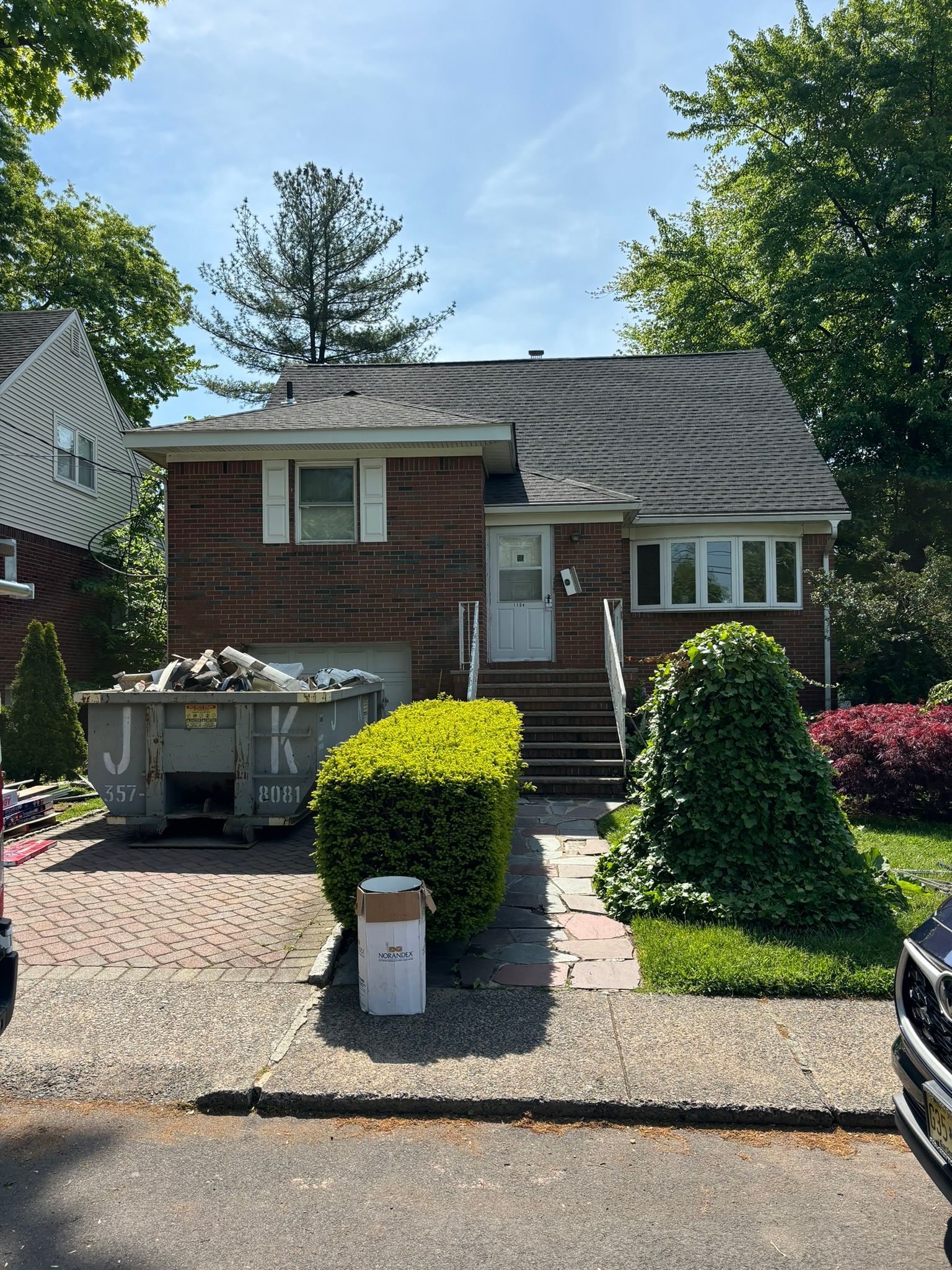 Brick house with small front yard, bushes, and a dumpster. Sunny day.