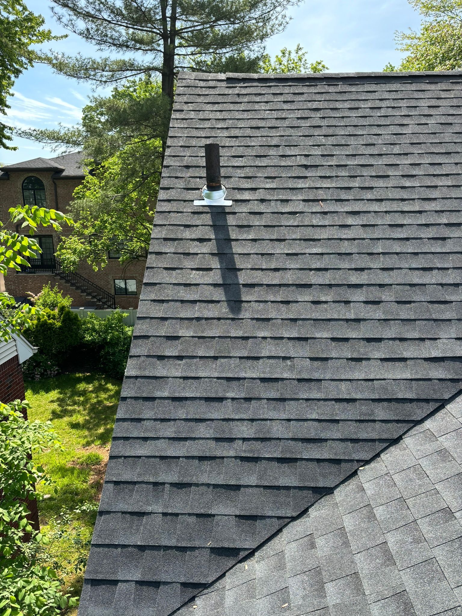 Dark gray shingle roof with a vent pipe and trees in the background. Sunny day.