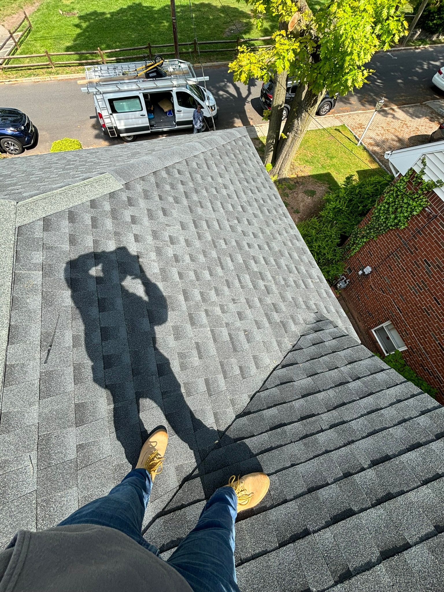 Person standing on a gray shingled roof, casting a shadow. A work van and trees are visible in the background.