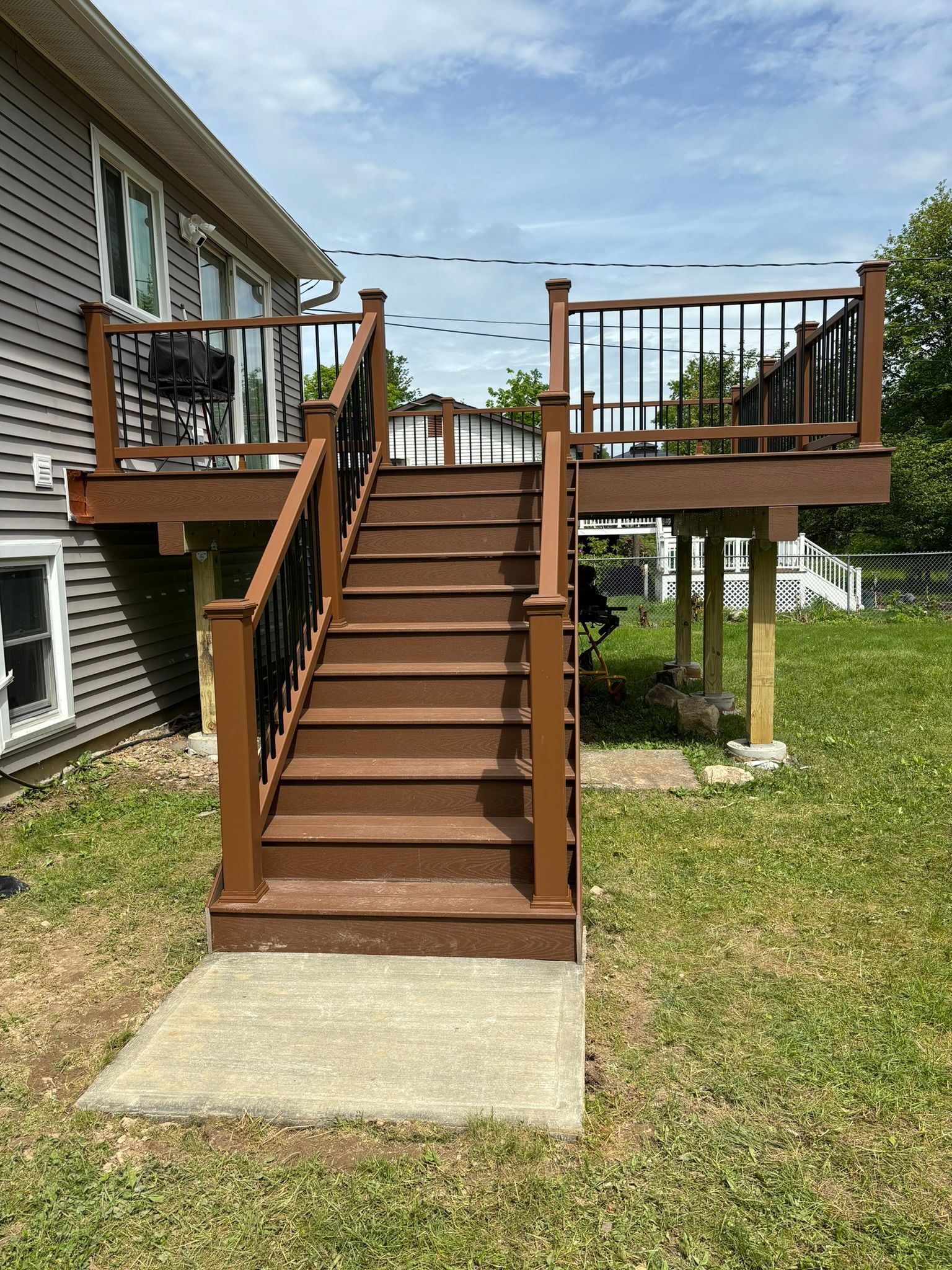 Brown deck with stairs, black railing, on a grassy lawn; house in background.