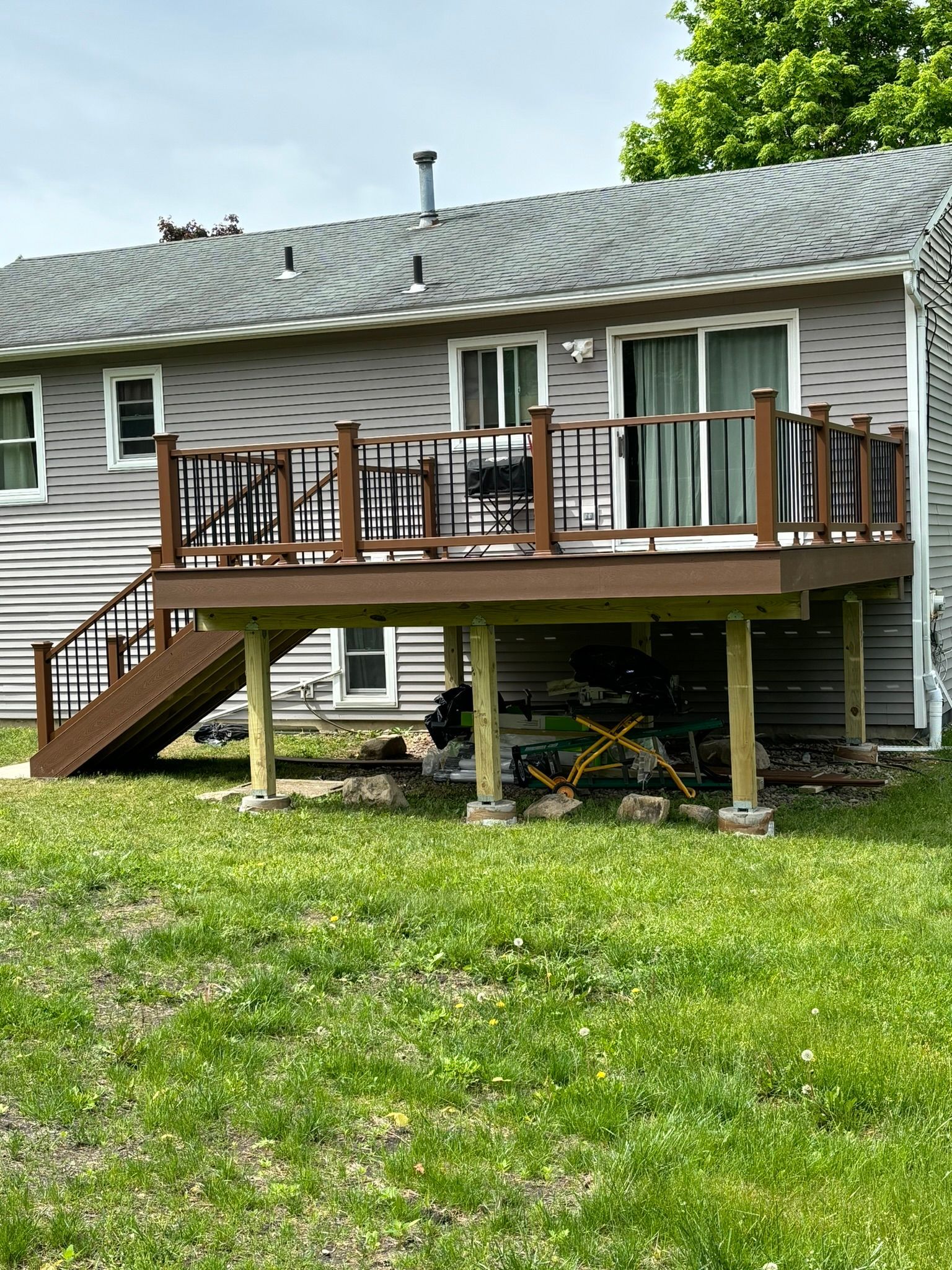 Brown deck with brown and black railing, stairs, and support posts attached to a grey house with a grassy lawn.