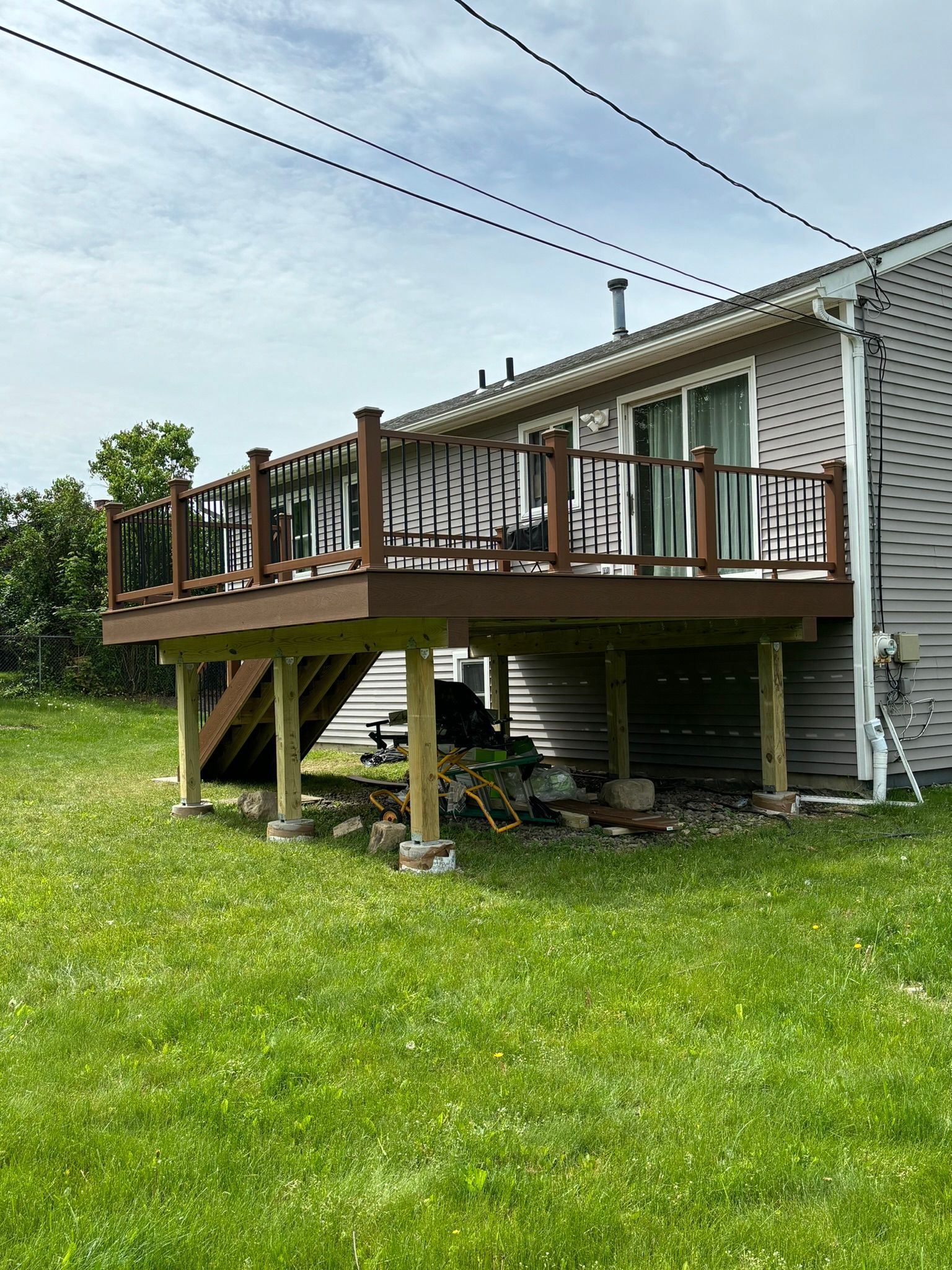 Elevated wooden deck attached to a house with black railings, brown posts, and a set of stairs on a grassy lawn.