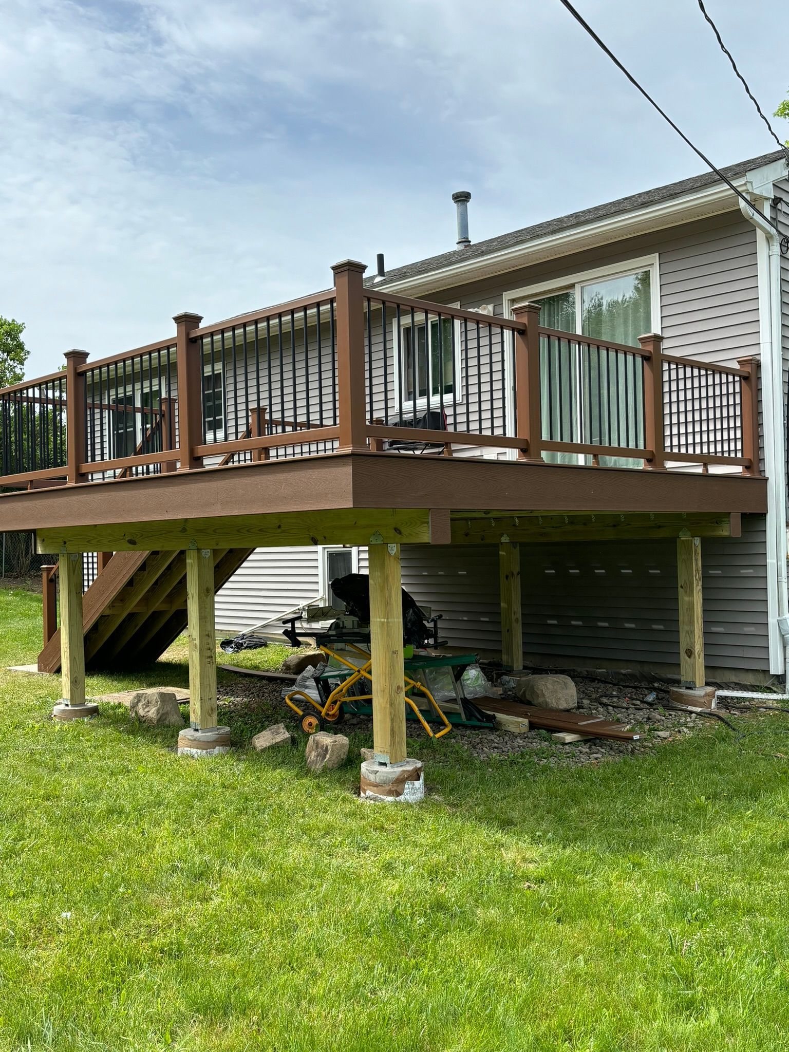 Elevated brown deck with black railing attached to a house; wooden support posts sit on concrete.