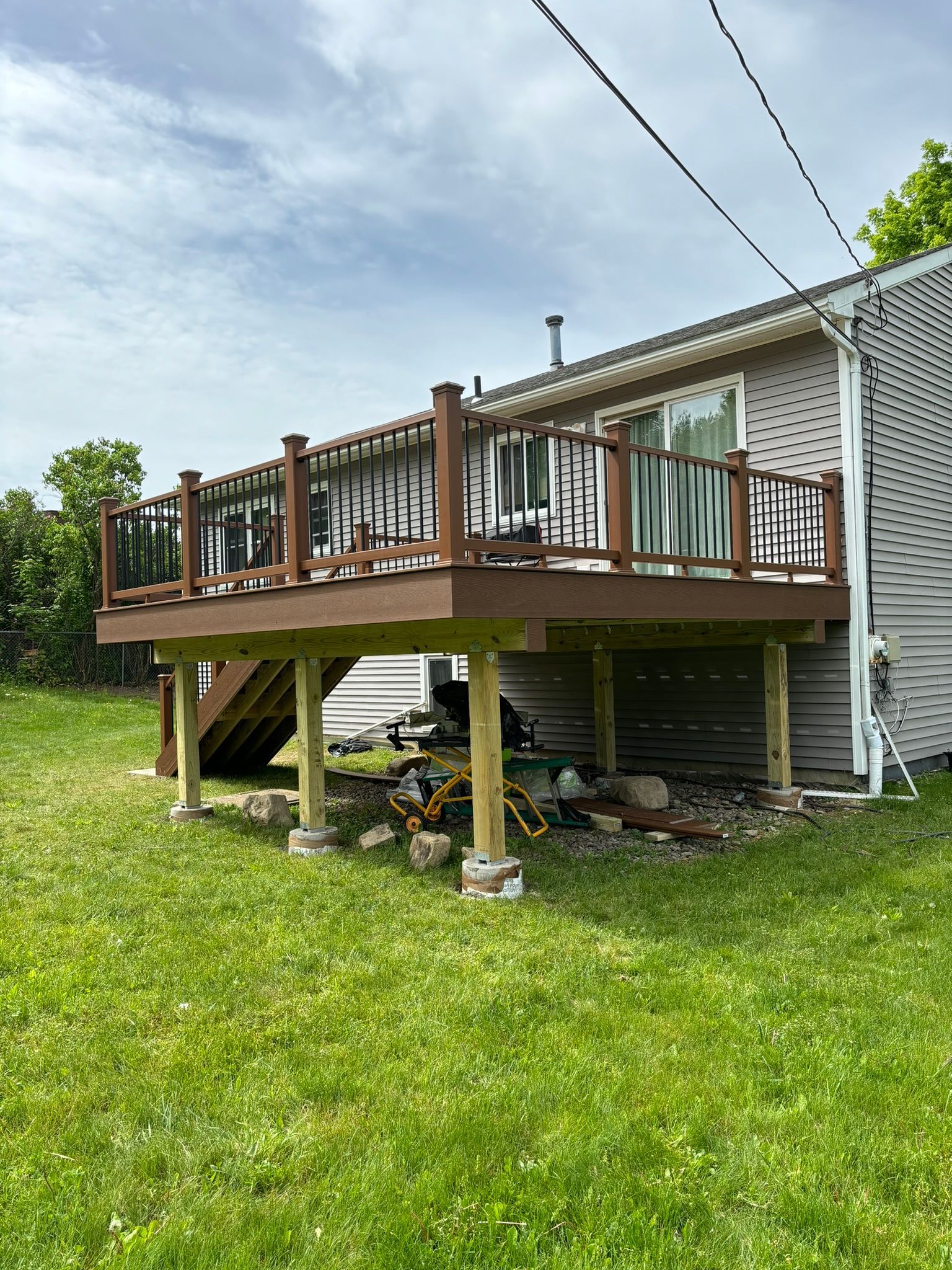 Elevated wooden deck attached to a house with railing, supported by posts on a grassy lawn.