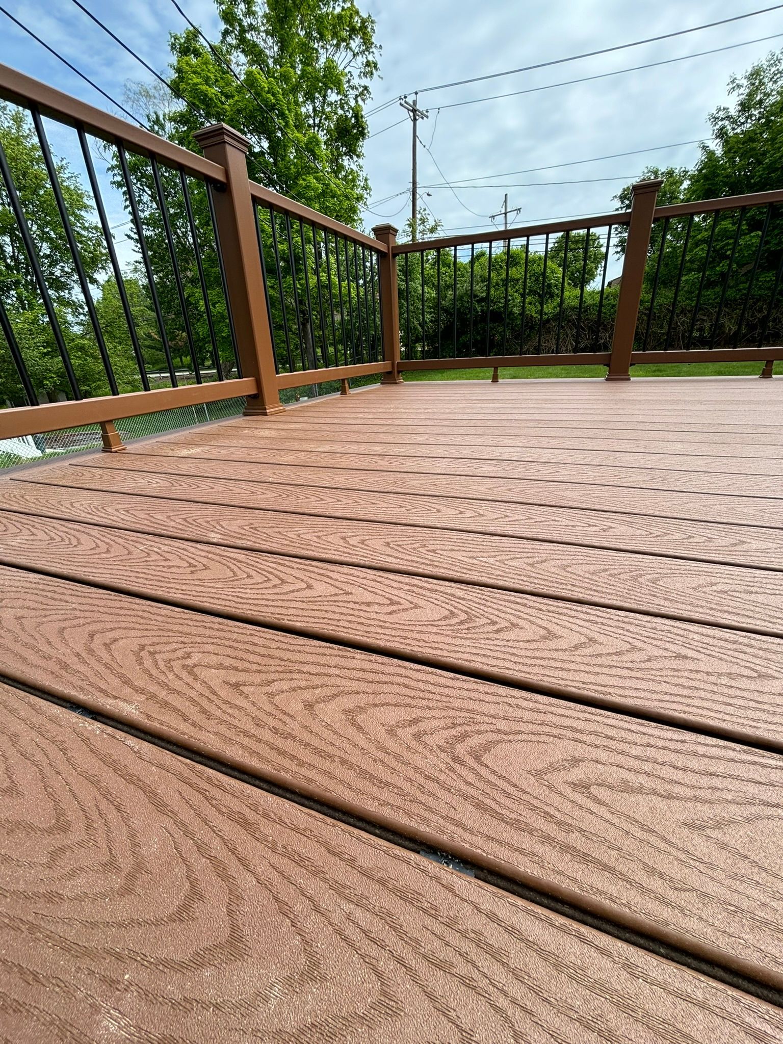 Brown composite deck with railing, overlooking green trees and a cloudy sky.