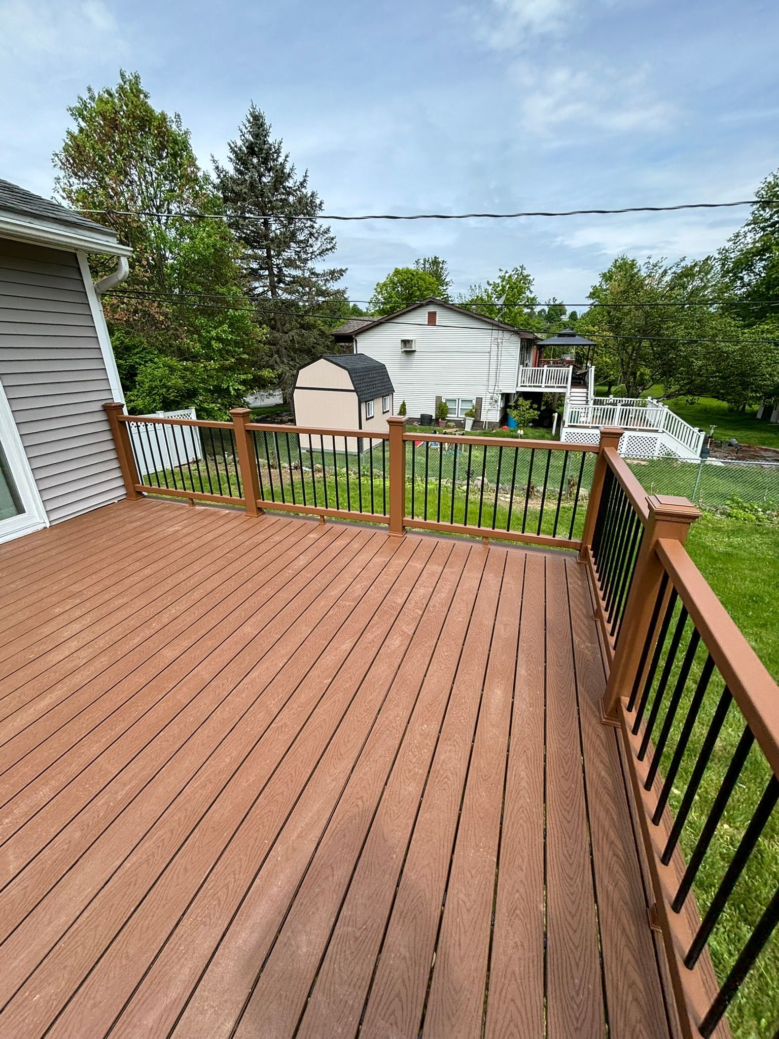 Wooden deck with brown planks and black railings, overlooking a backyard with houses and trees.