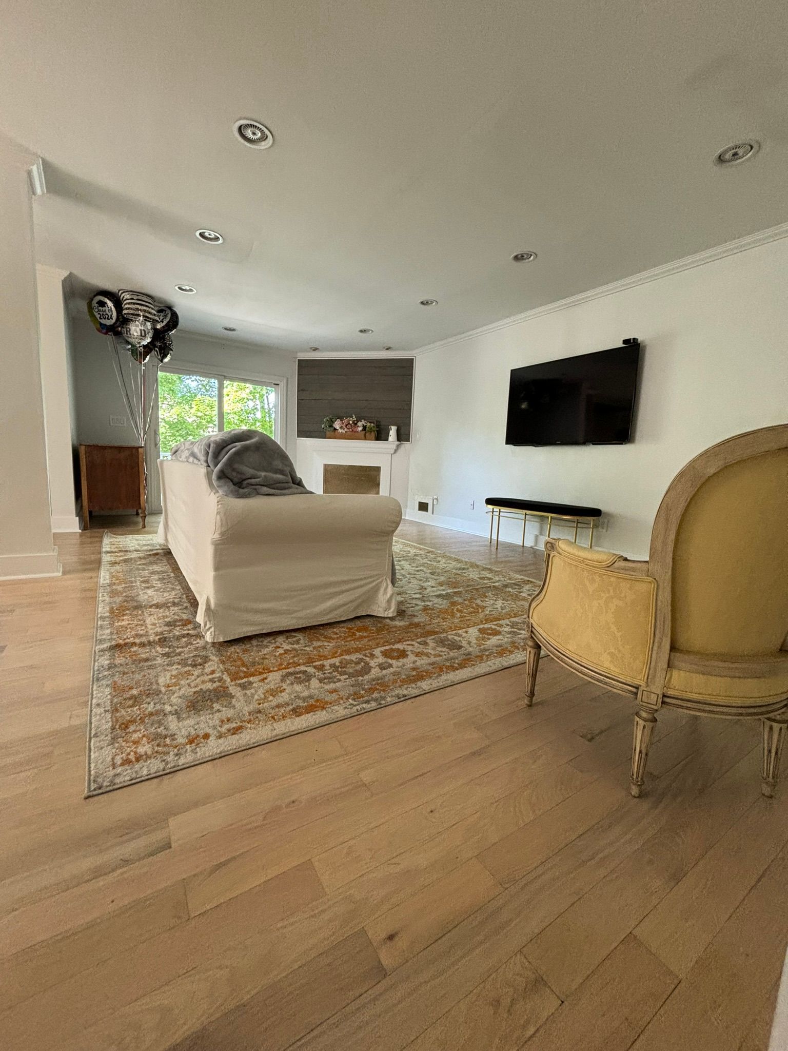 Living room with light wood floors, rug, sofa, fireplace, and TV on the wall. A yellow armchair is in the foreground.