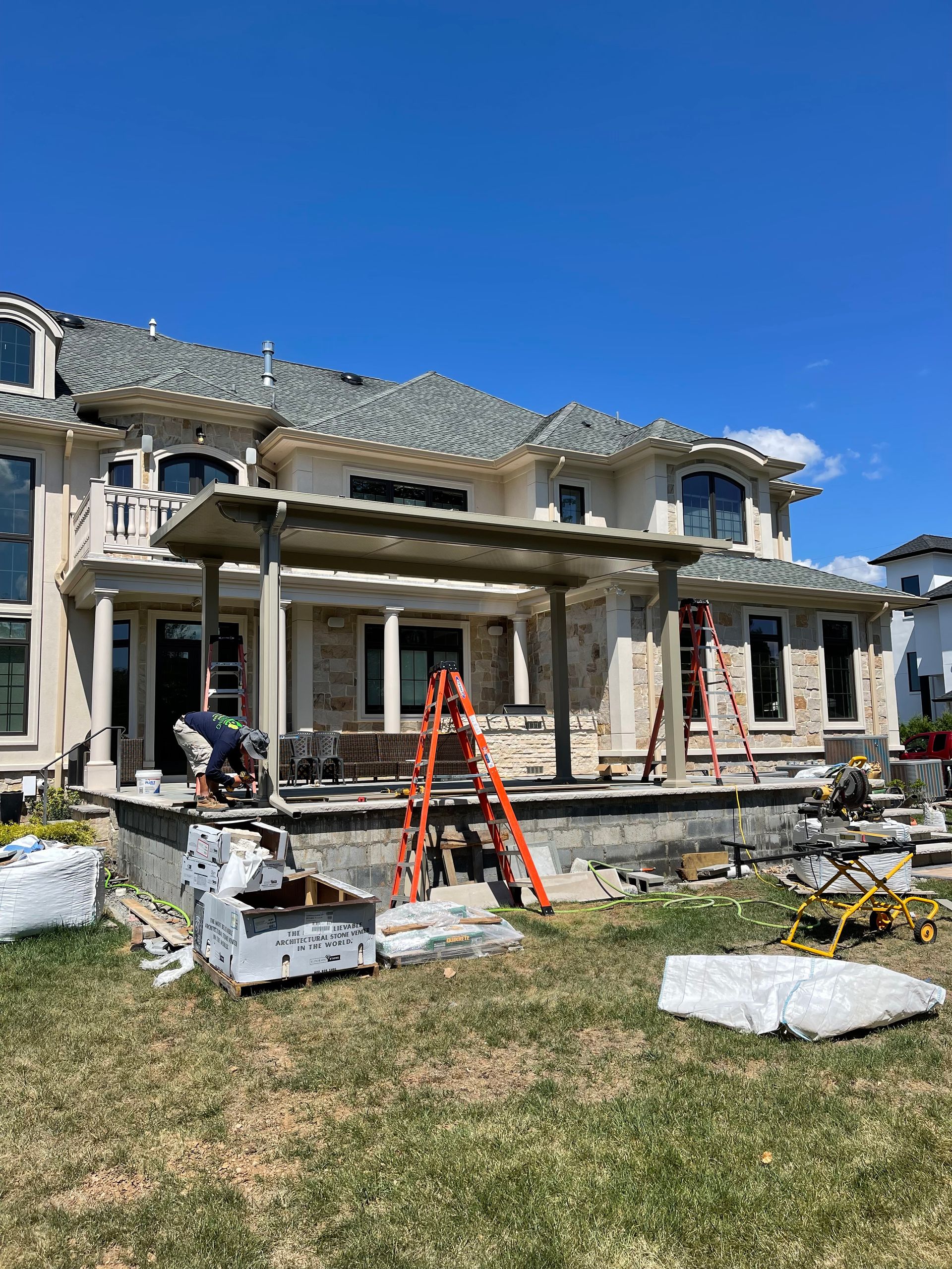 Construction on a large house with a stone patio. A worker is seen with ladders and tools. Blue sky.