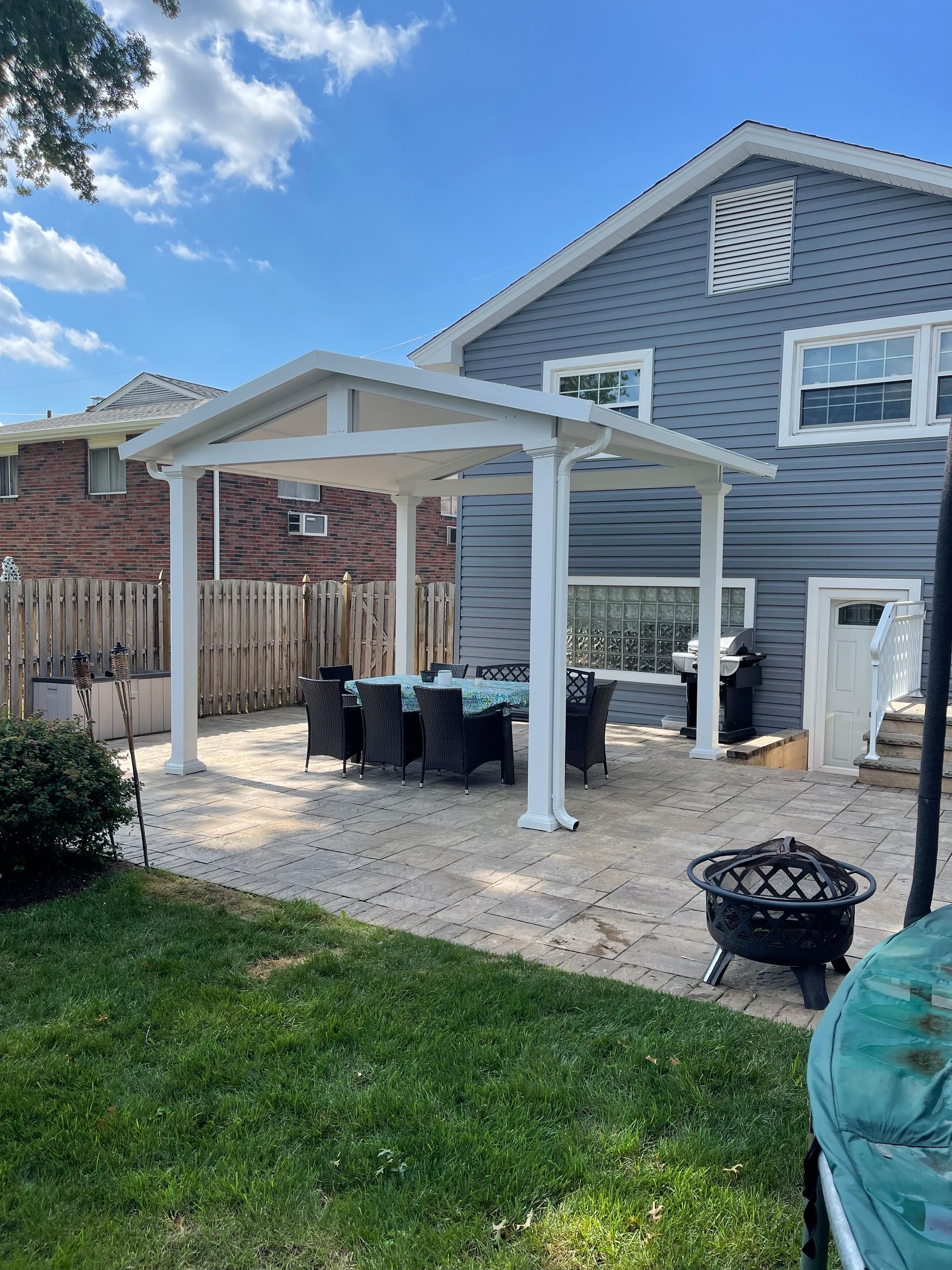 Backyard patio with a white pergola, table, and chairs next to a blue house on a sunny day.