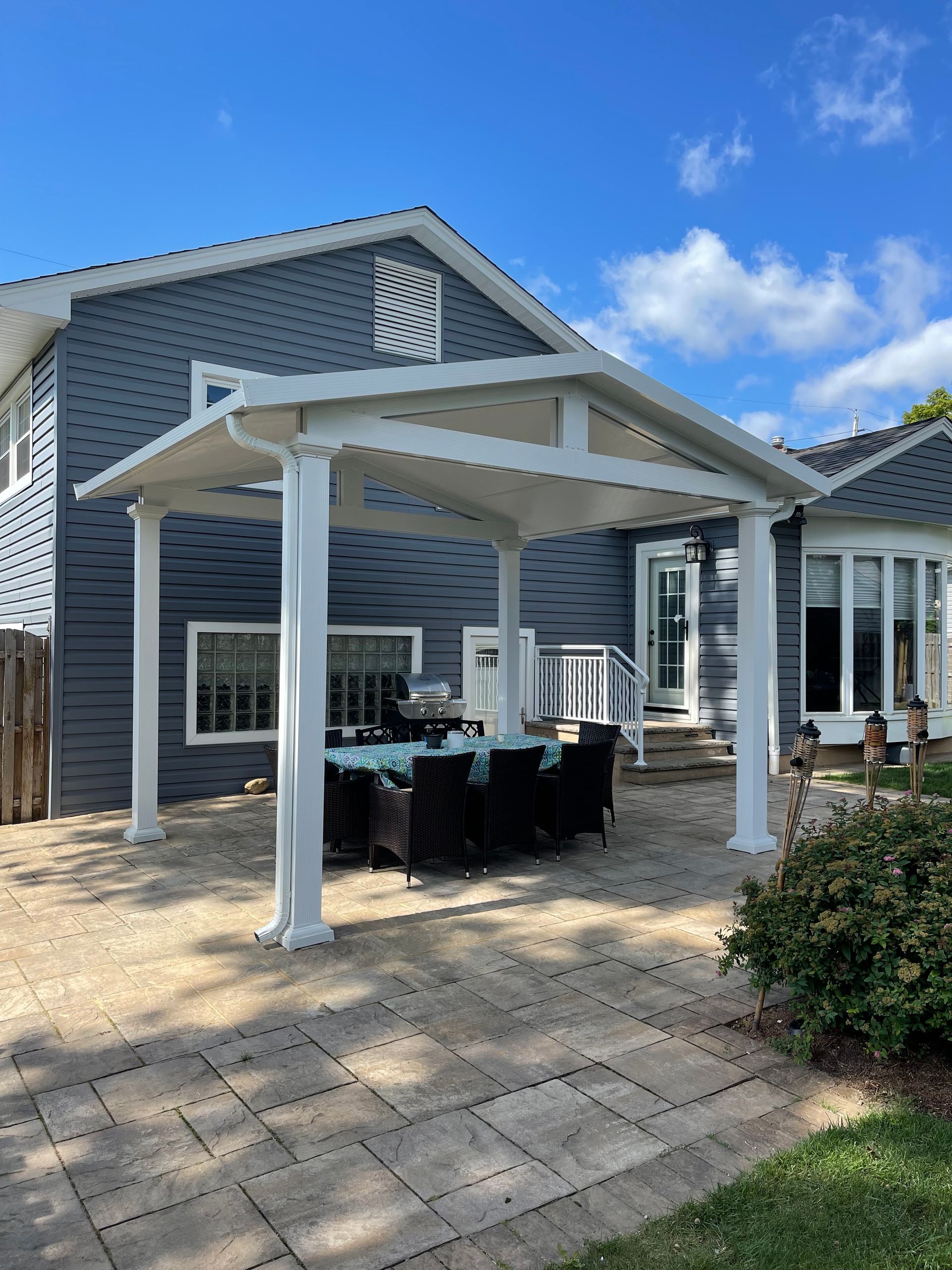 White-framed patio cover over outdoor dining set on a stone patio, adjacent to a blue house on a sunny day.