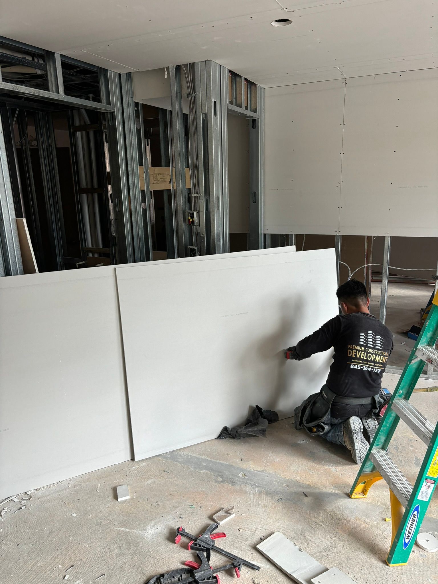 Construction worker installing drywall in a room with metal framing; a ladder and tools are visible.