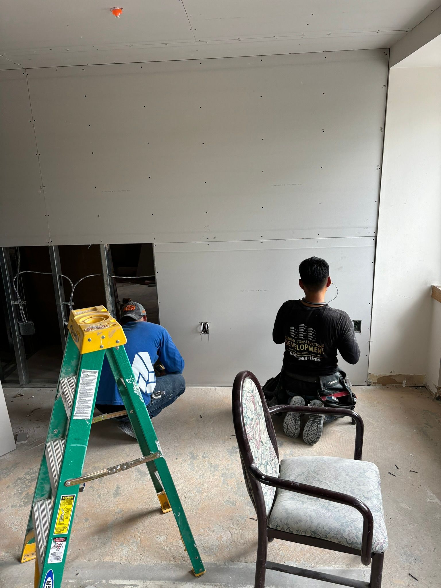 Two people installing drywall in a room. One kneels, the other sits on a chair. Green ladder nearby.
