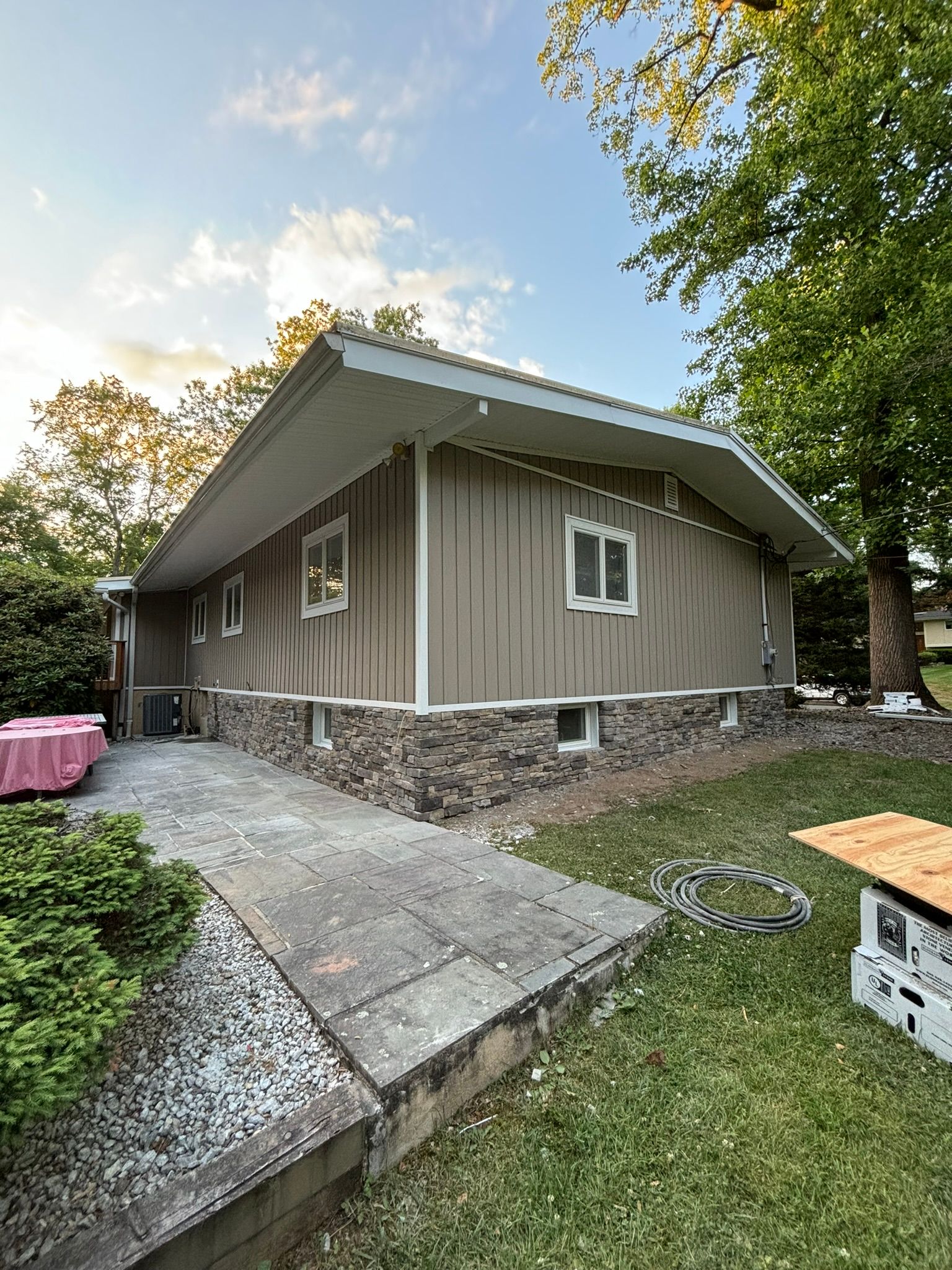 Tan-sided house with stone base. Two windows, white trim, gray roof, pathway. Green yard with tree and bushes.