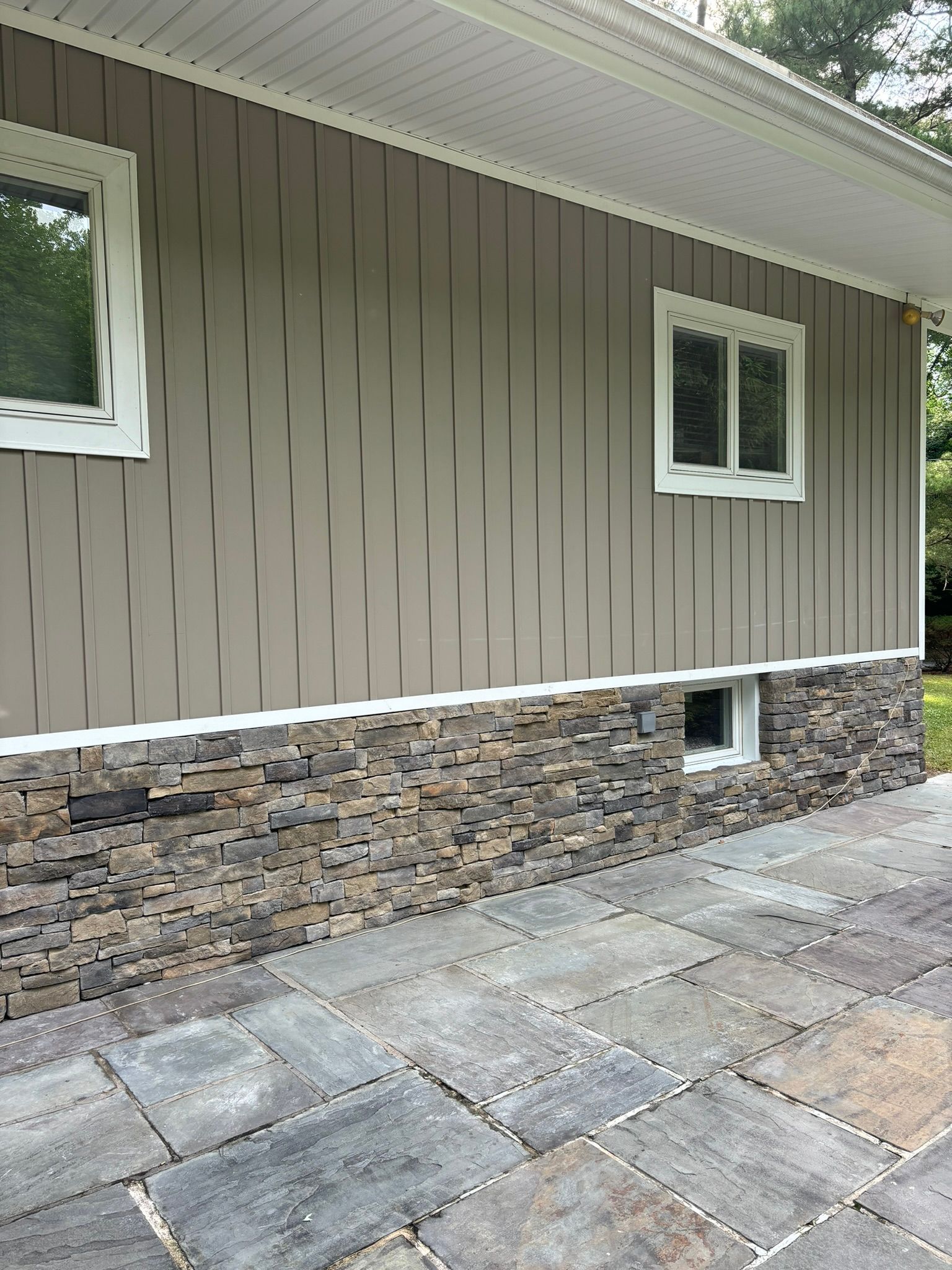 Tan siding and stone facade on a house exterior with two windows and a paved patio.