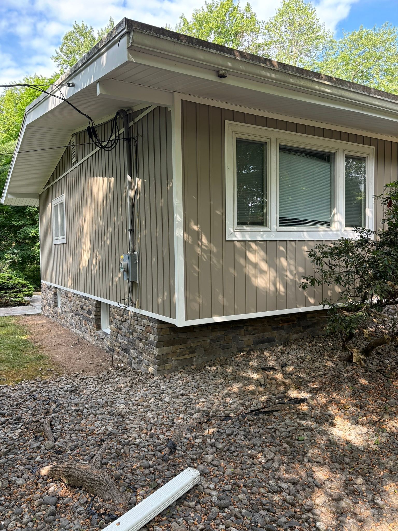Tan house with white trim and a stone foundation, set on a gravel slope.