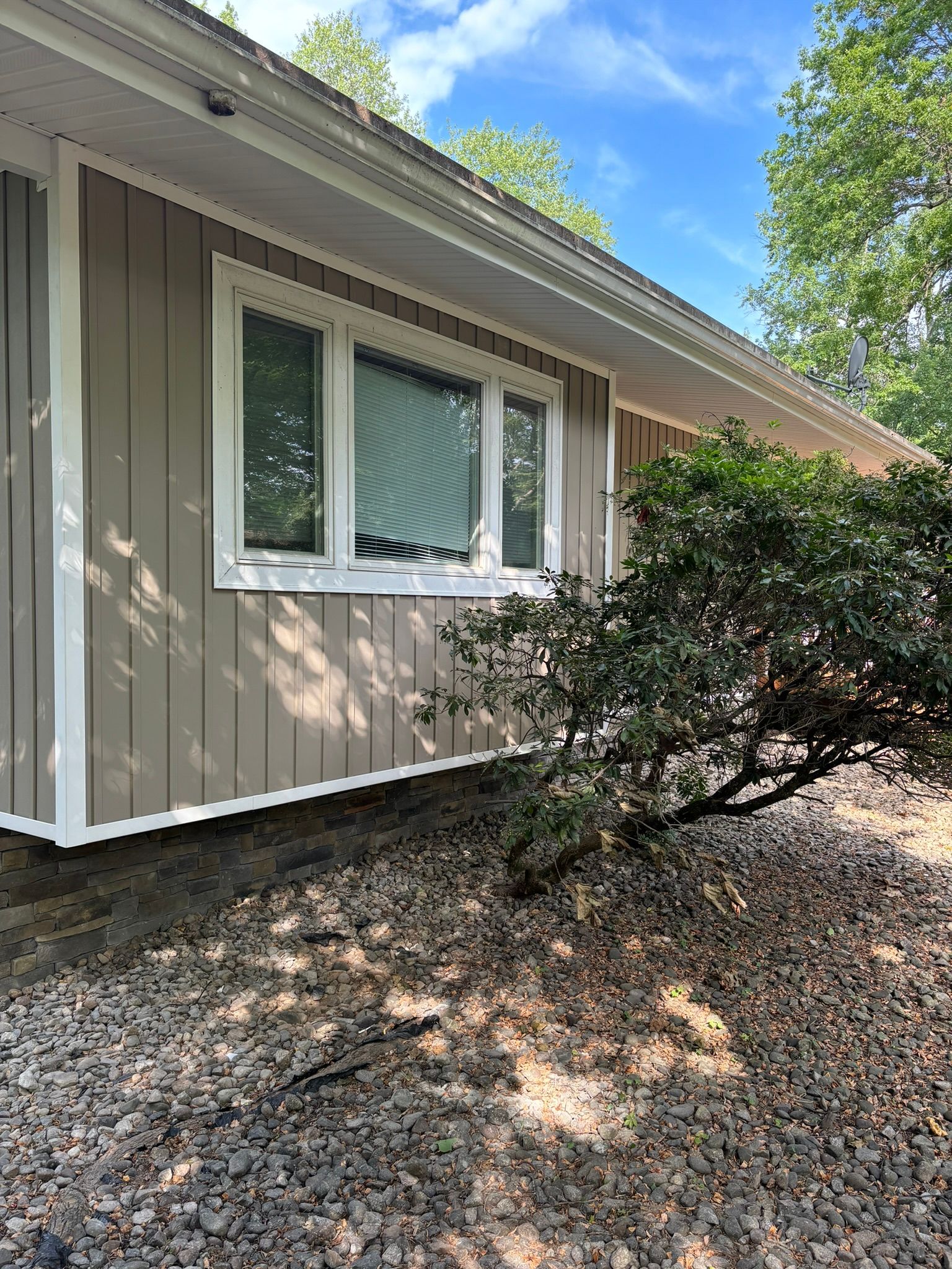 Tan siding on a house with white trim. Window with closed blinds. Green bush and rocky ground in front.