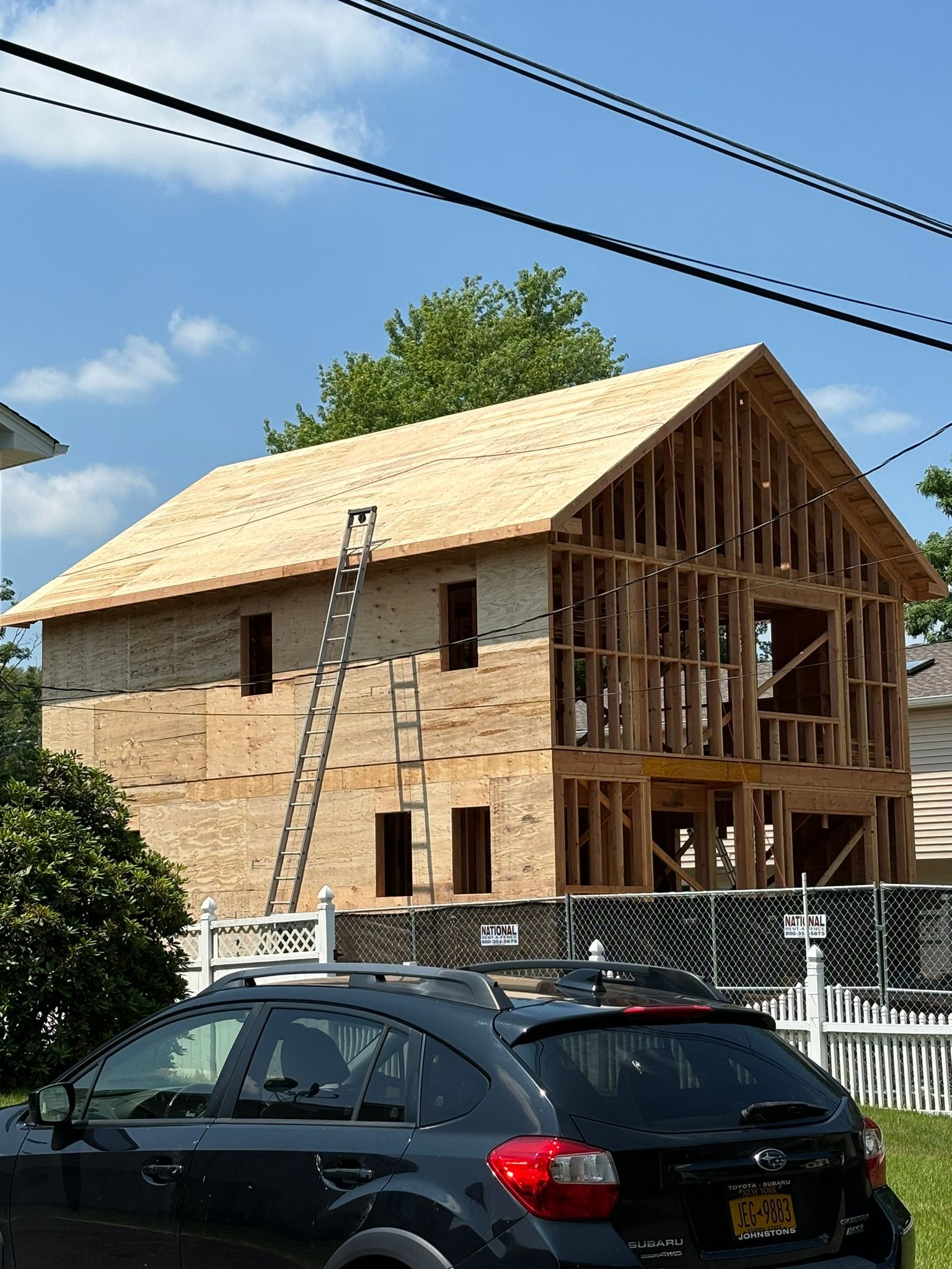 Two-story house under construction; wooden frame, plywood siding, ladder against the side, car in front.