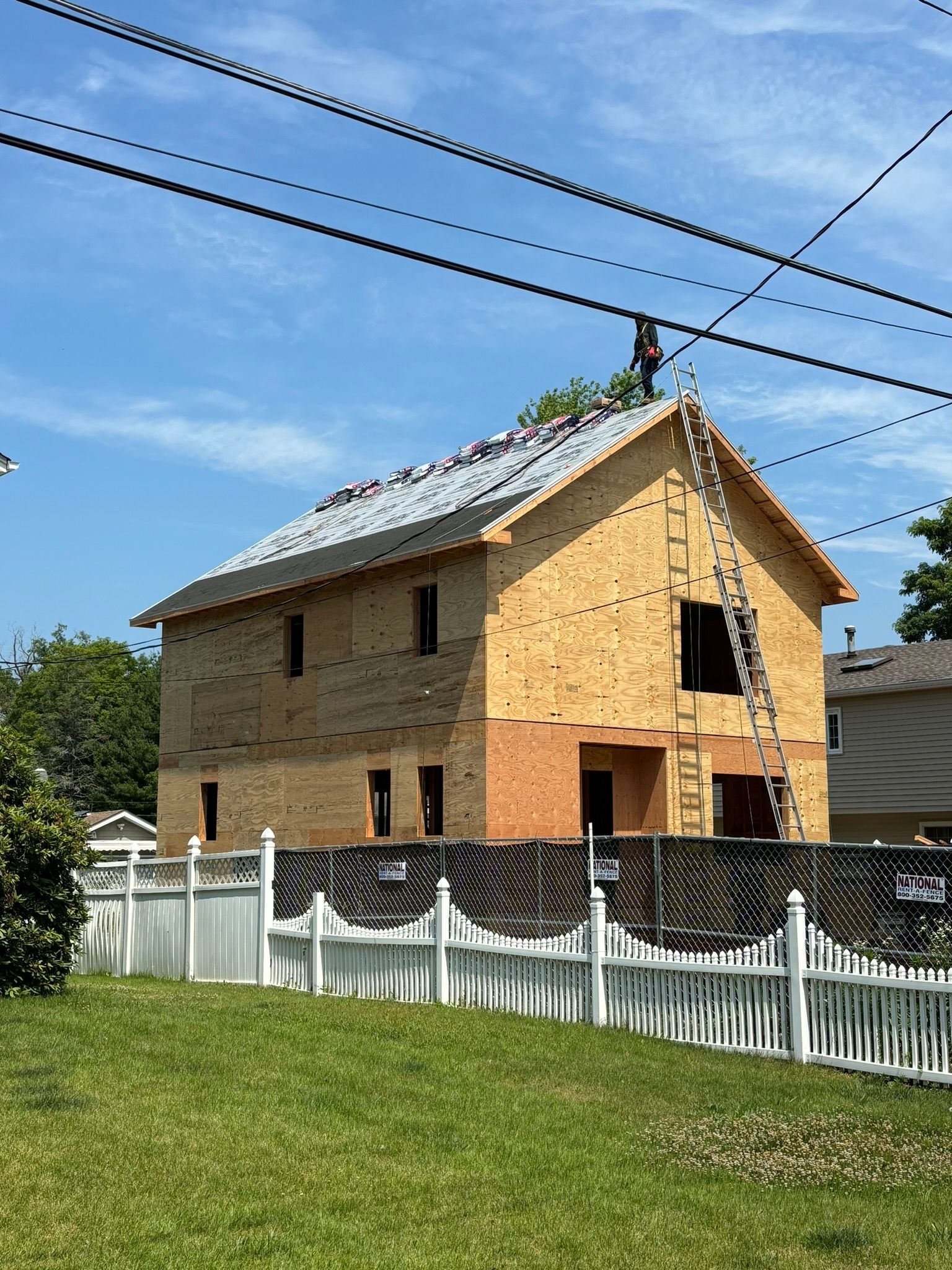 Two-story house under construction, framed with plywood, ladder, and a partially built roof. White fence in foreground.