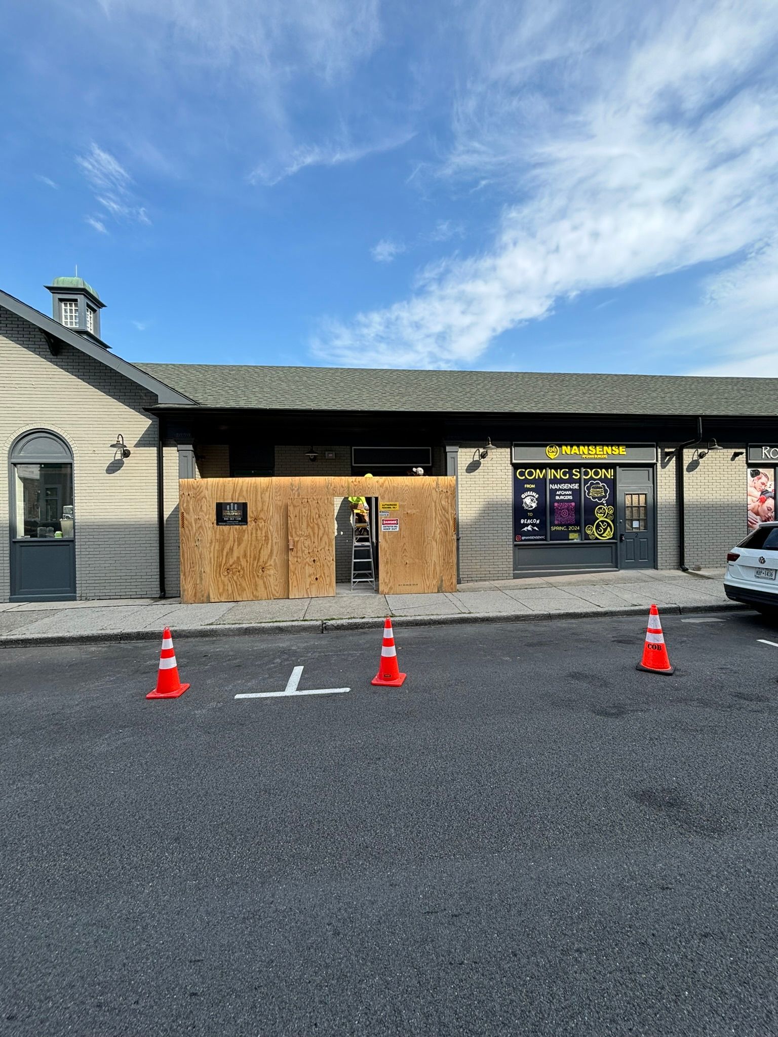 Exterior view of a building with a boarded-up entrance. Orange traffic cones and white lines mark the area.