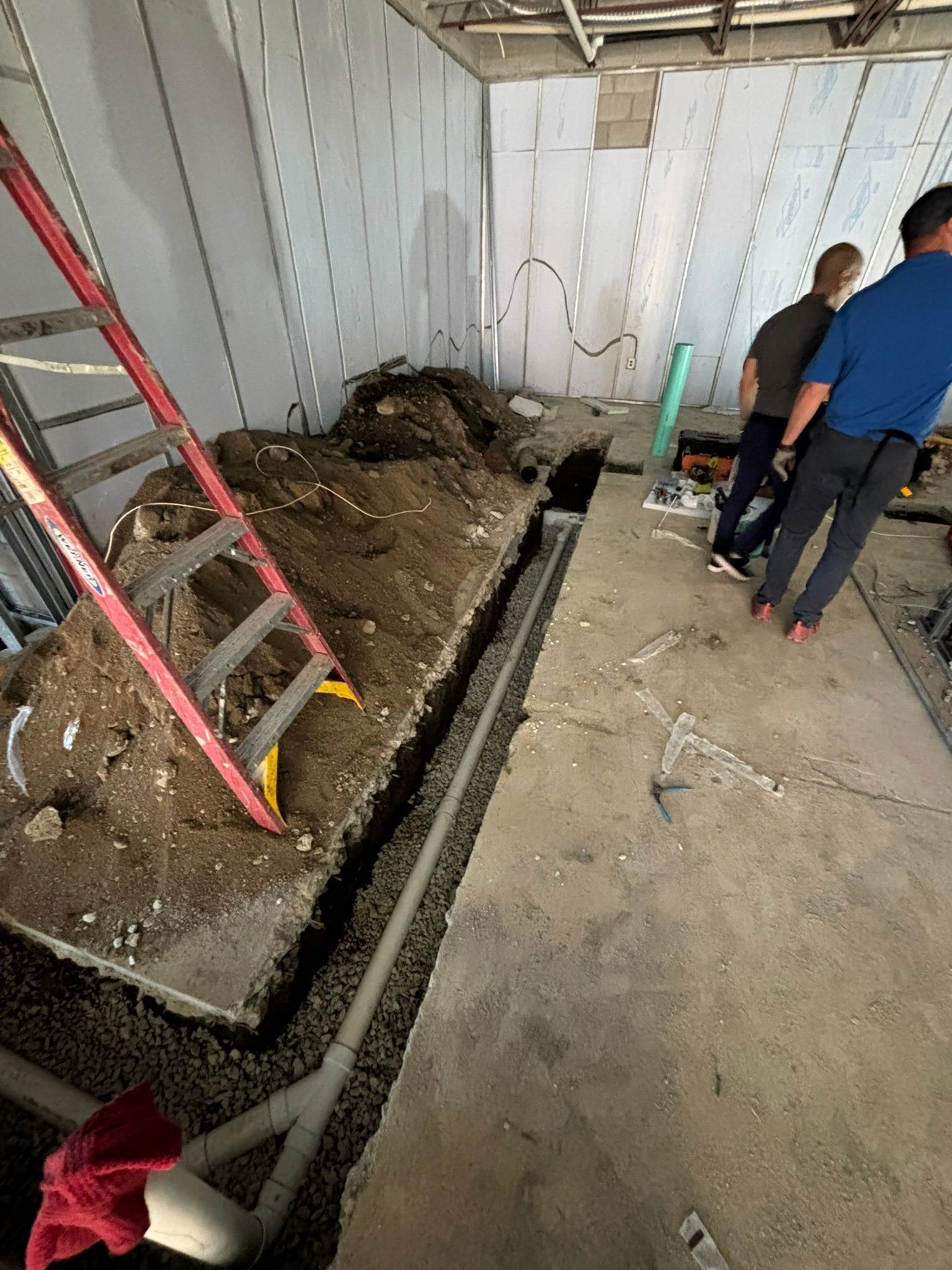 Construction site: trench dug with pipe laid. Workers near unfinished walls and debris.