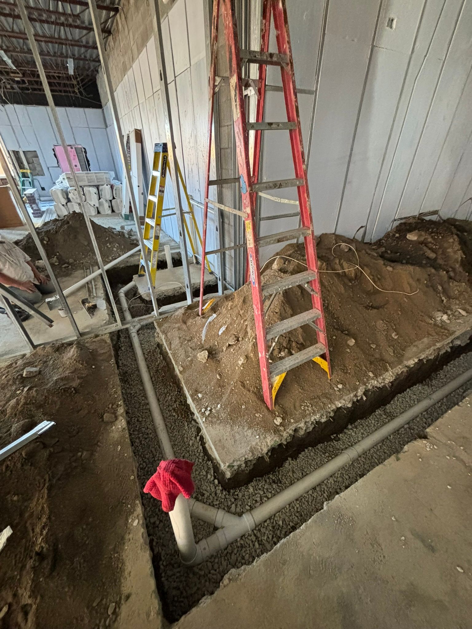 Construction site with red ladder, plumbing, dirt, and gravel. Interior with unfinished walls.
