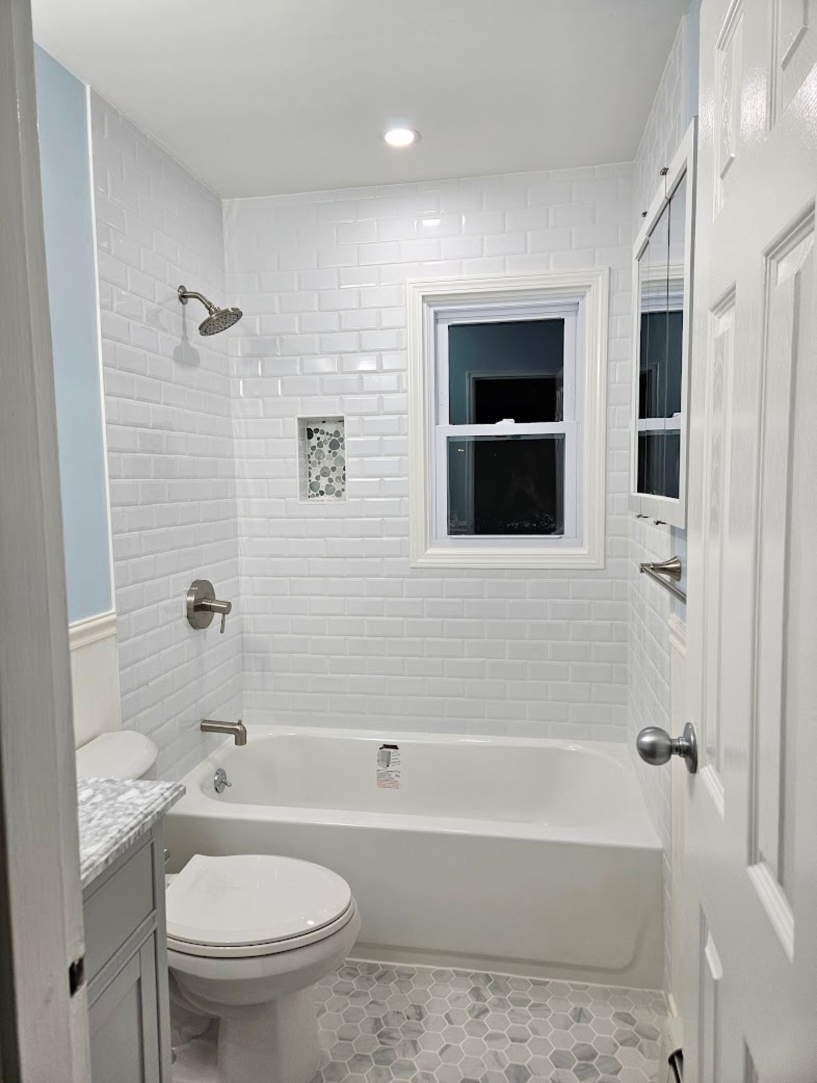 White tiled bathroom with tub, toilet, window, and small pebble flooring.