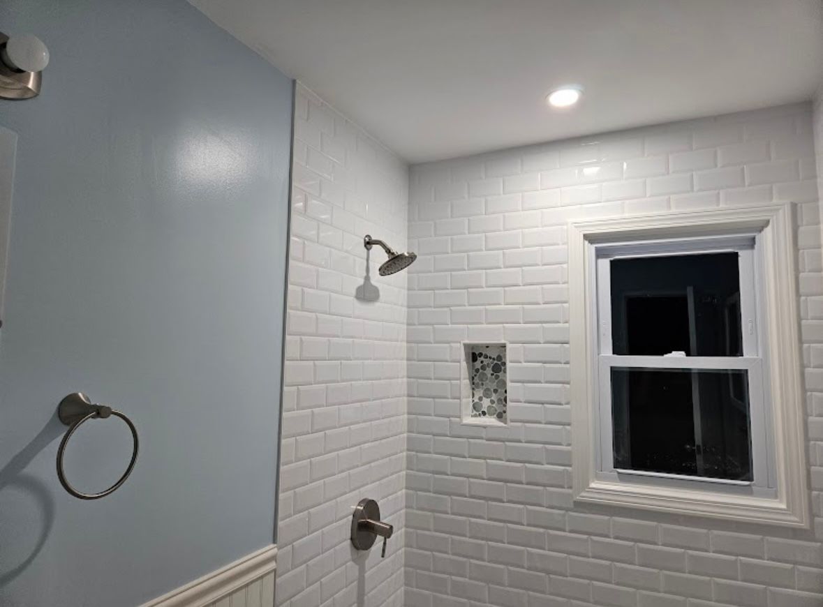 White tiled shower with a small window, light blue wall, and chrome fixtures.