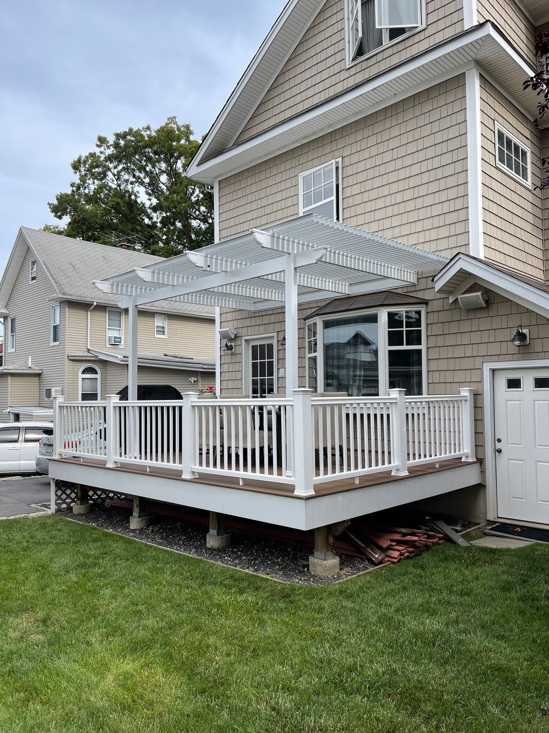 White pergola over a deck with white railing, attached to a beige house, with green grass in the foreground.