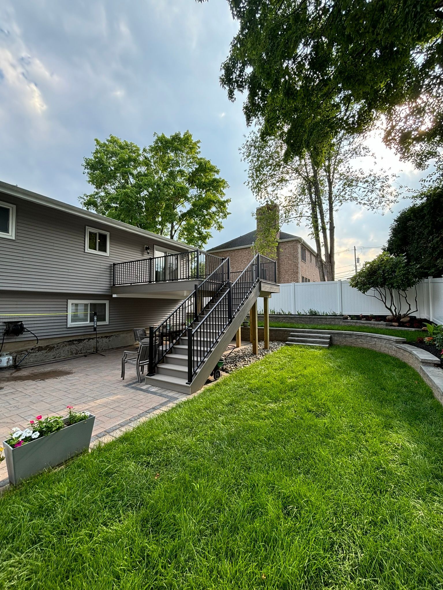 Backyard with deck, stairs, and retaining wall. Gray house, black railing, green grass, stone patio, and trees.