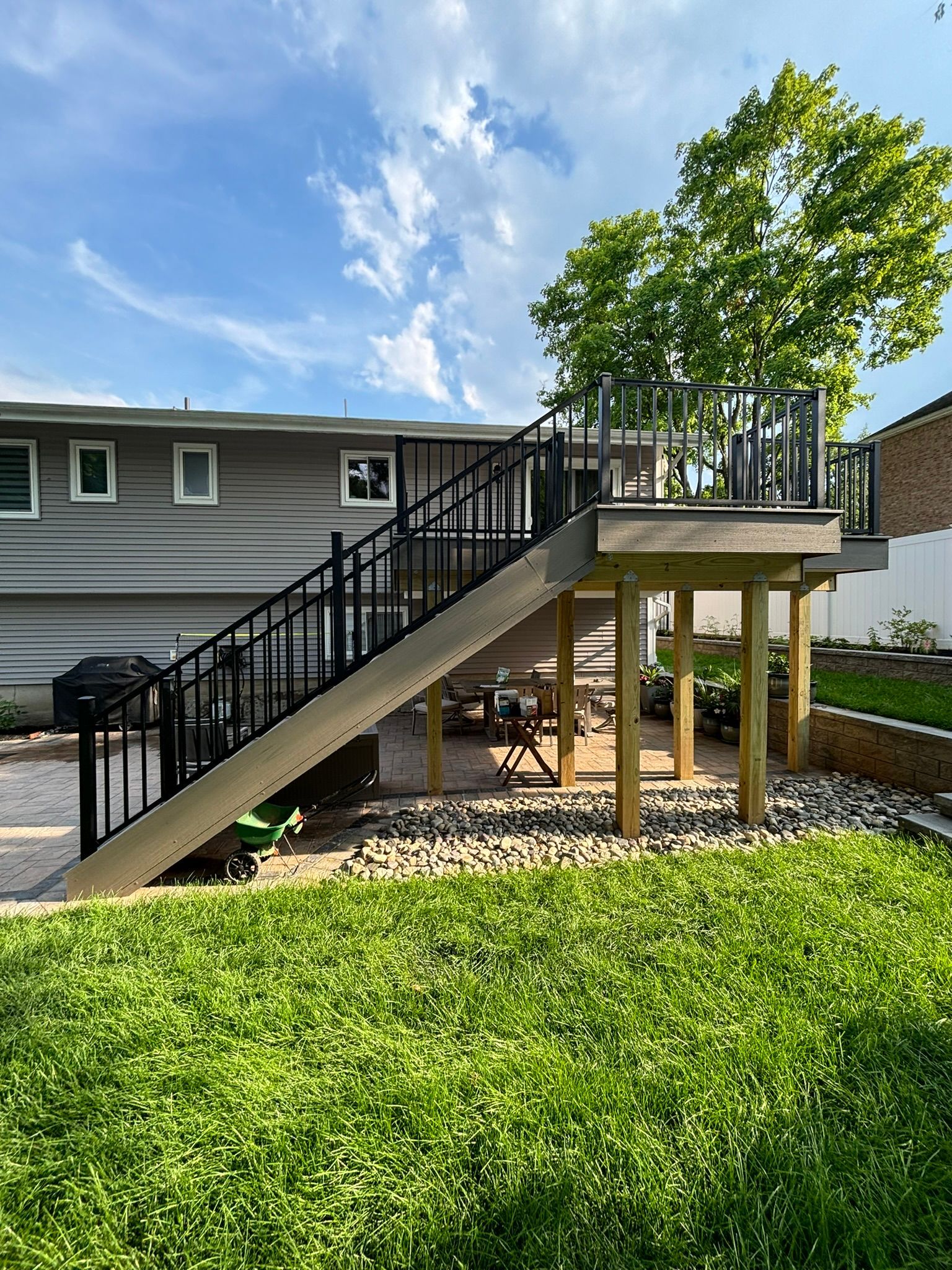 A raised wooden deck with black railings and stairs leading to a backyard with green grass and a gravel bed.