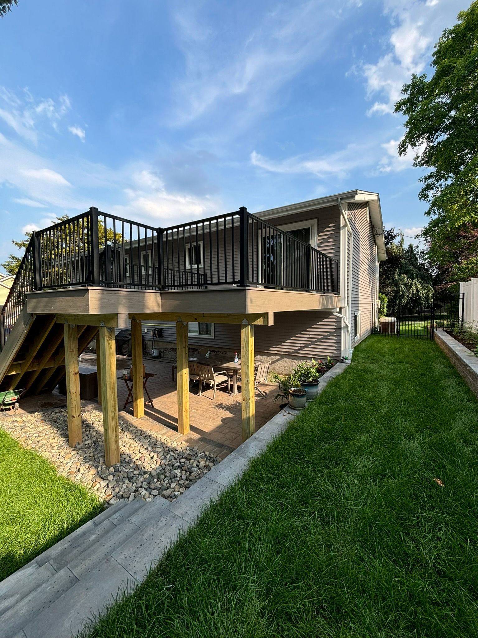 Elevated wooden deck with black railing and stairs next to a house with a grassy lawn and blue sky.