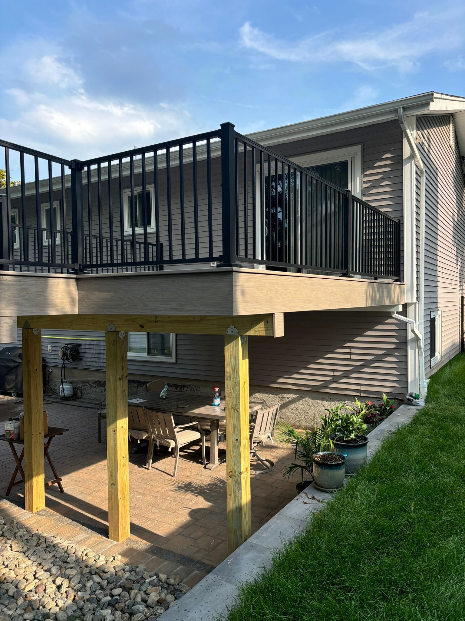 Deck with black railing, supported by wooden posts, overlooking a patio with furniture, next to a house with grey siding.