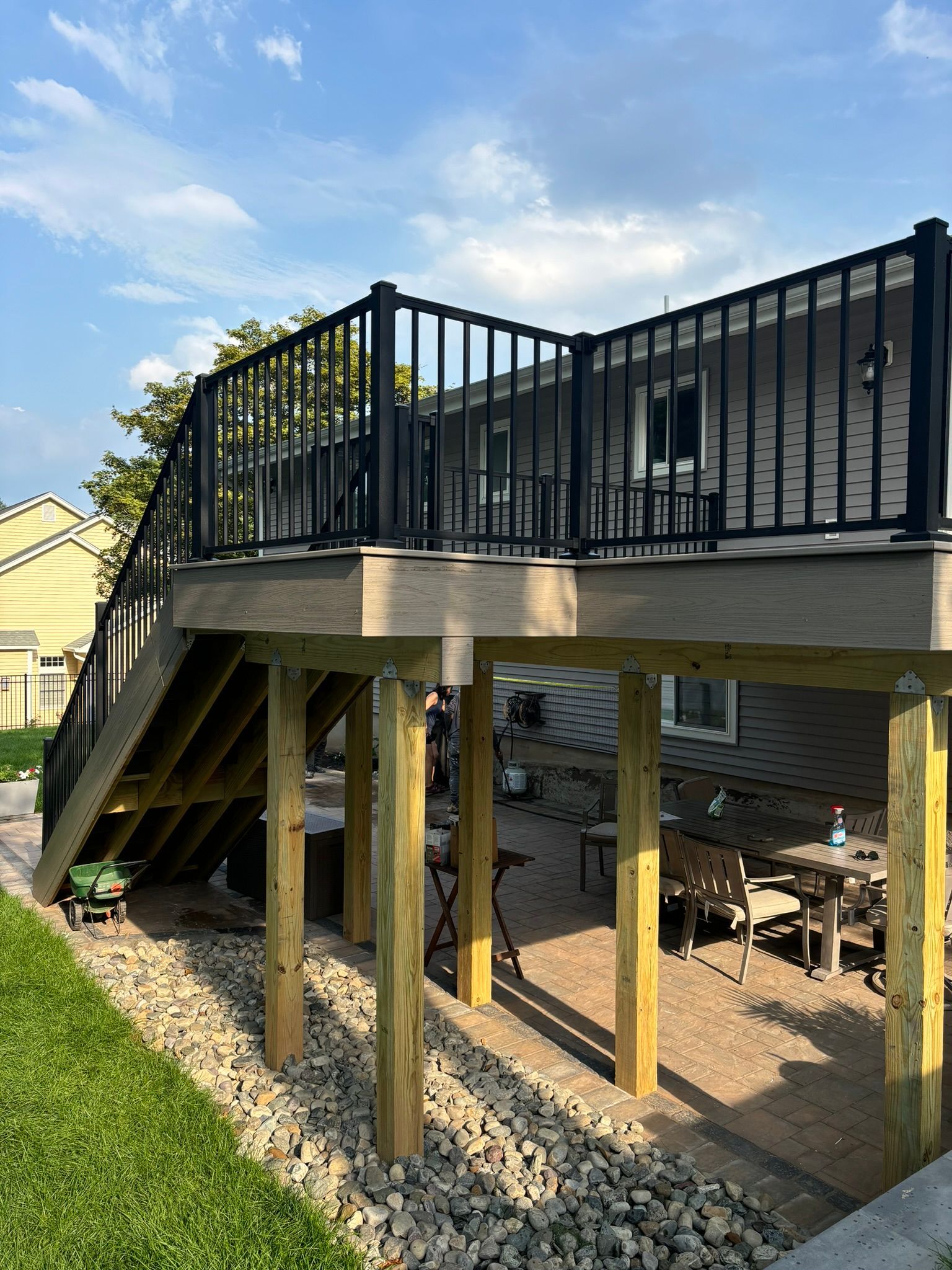 Elevated wooden deck with black railings and stairs. Underneath is a patio with tables, chairs, and a gravel base.