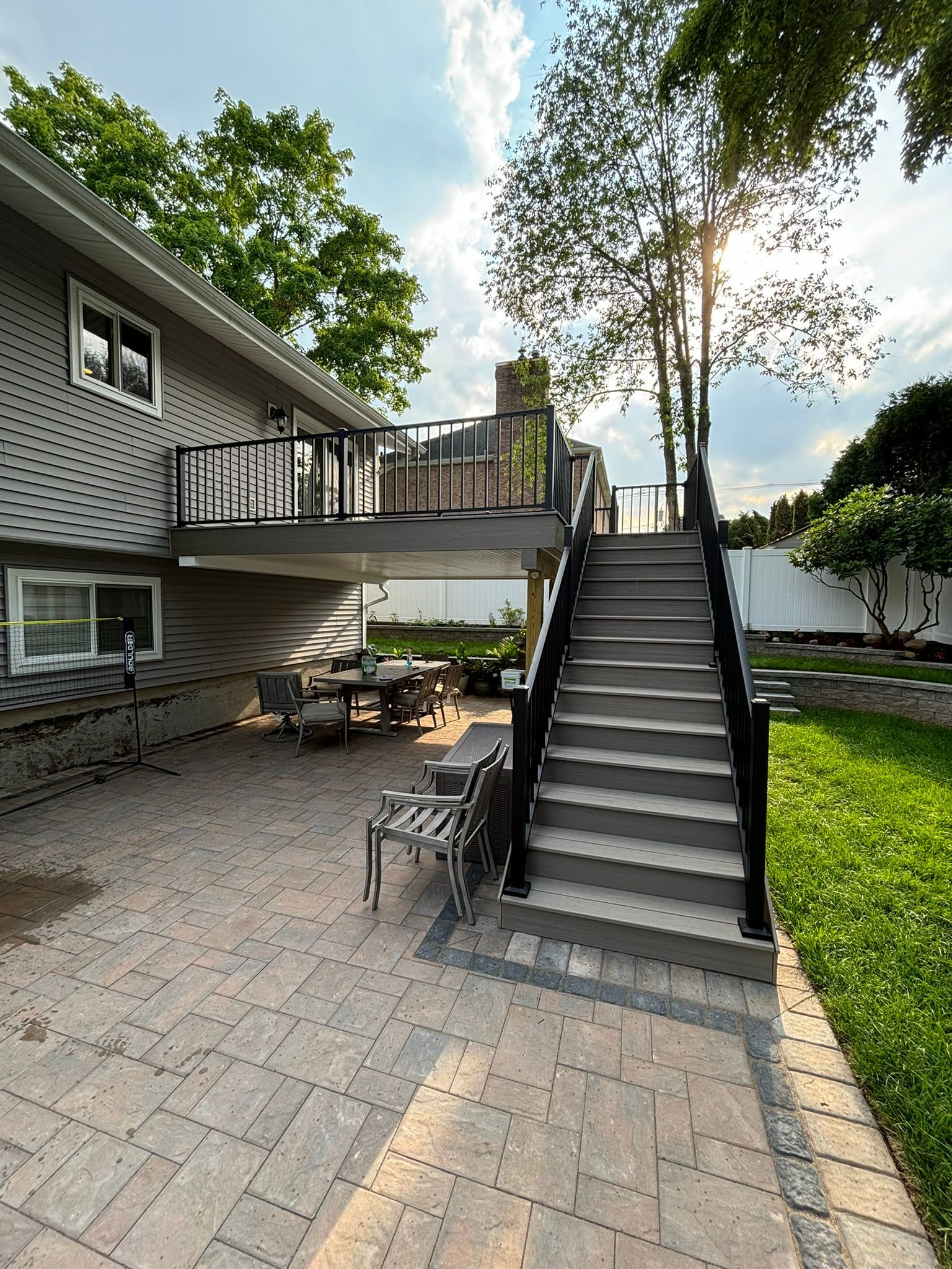Backyard with a deck, stairs, patio, and seating. Gray house, brick patio, green grass, trees, and bright sky.