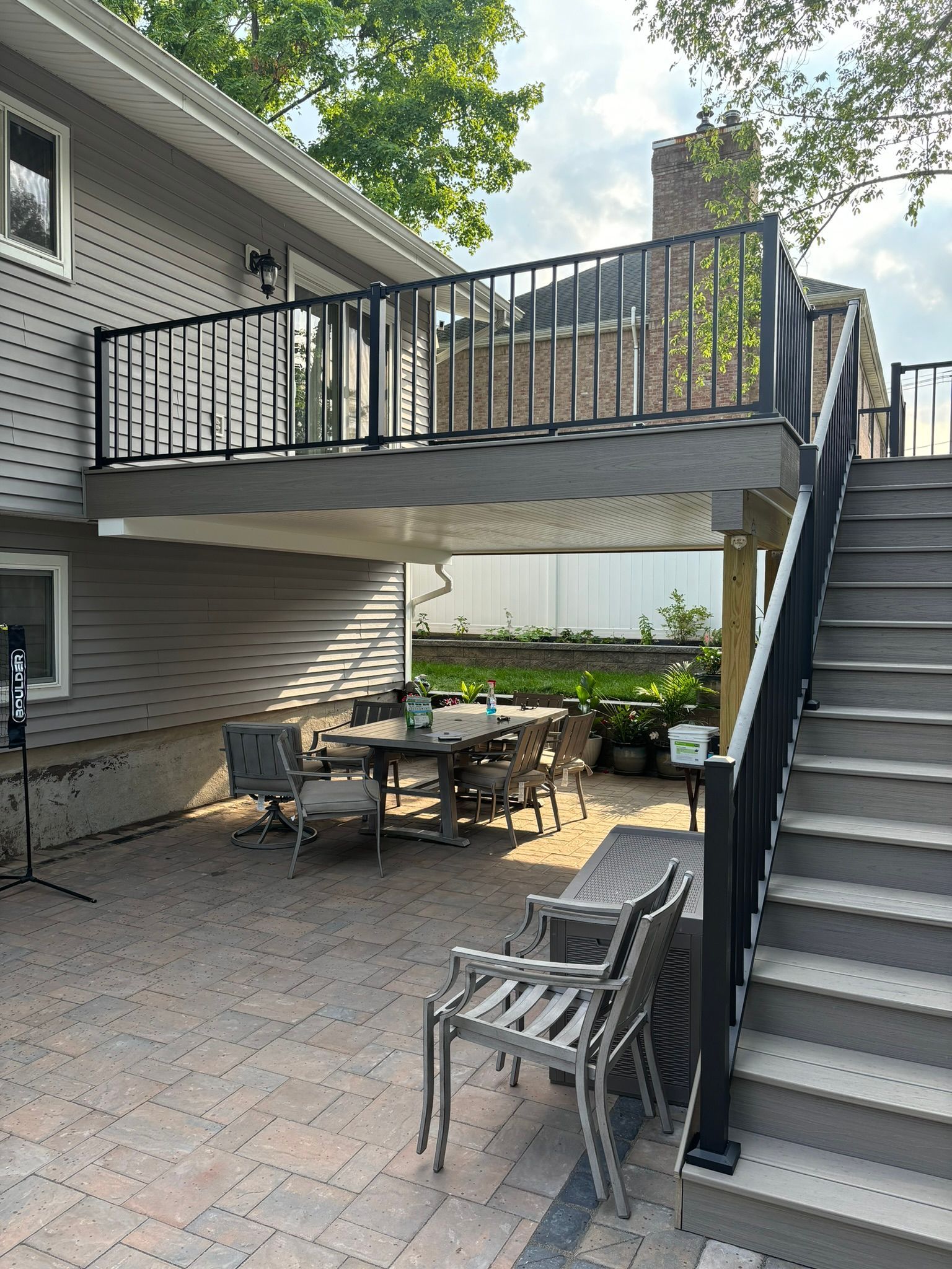 Patio under deck with stairs, dining set, and metal chairs. Gray siding, black railing, and brick pavers.