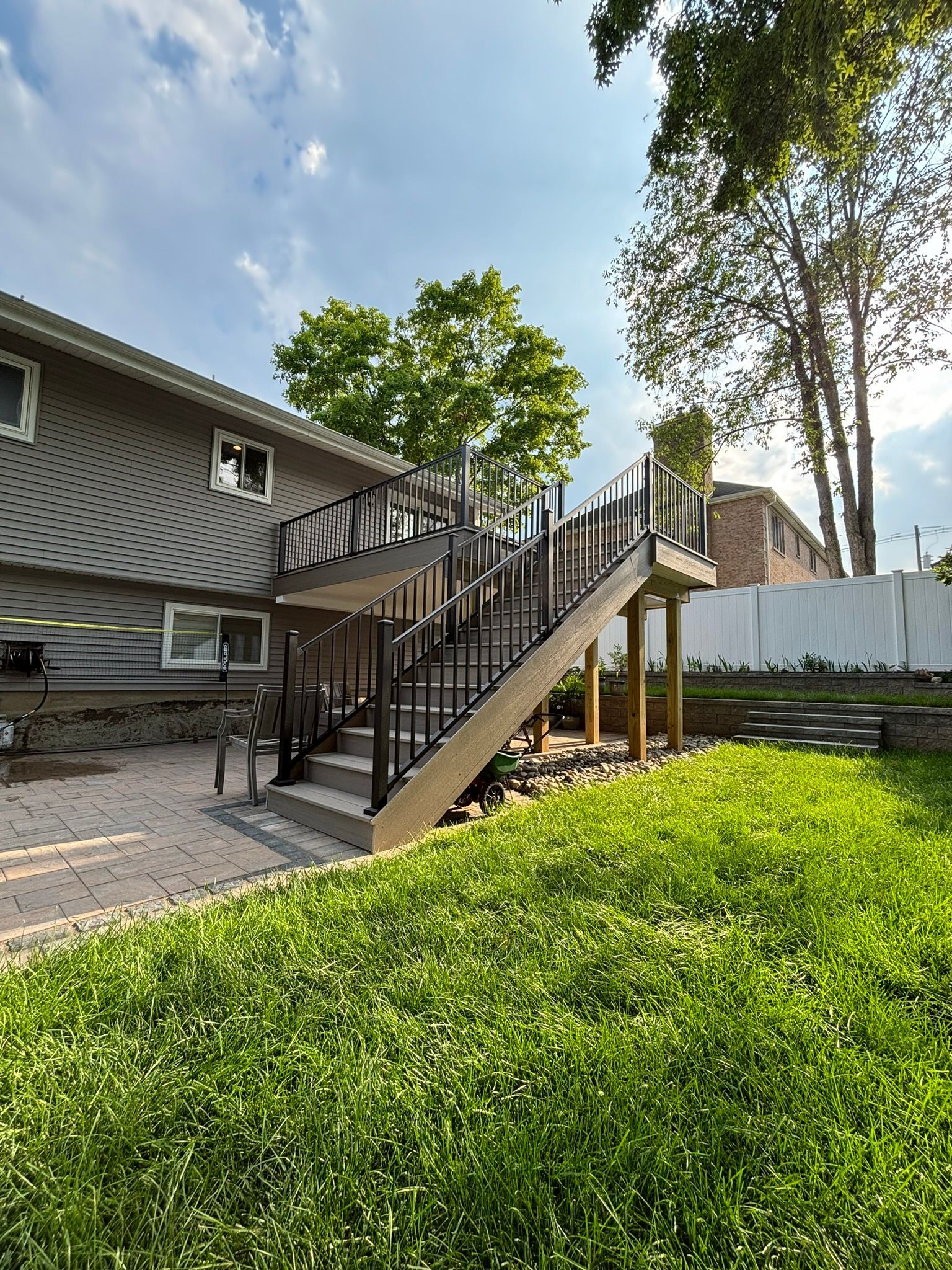 Backyard with a gray house, wooden deck with stairs, and a green lawn.