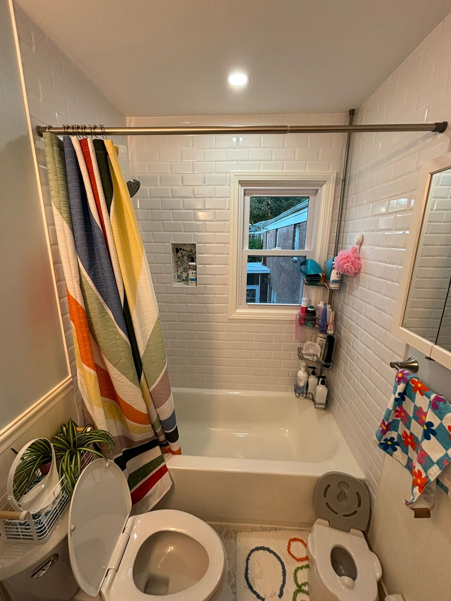 Small bathroom with tub, toilet, and window, white tile walls, colorful shower curtain.